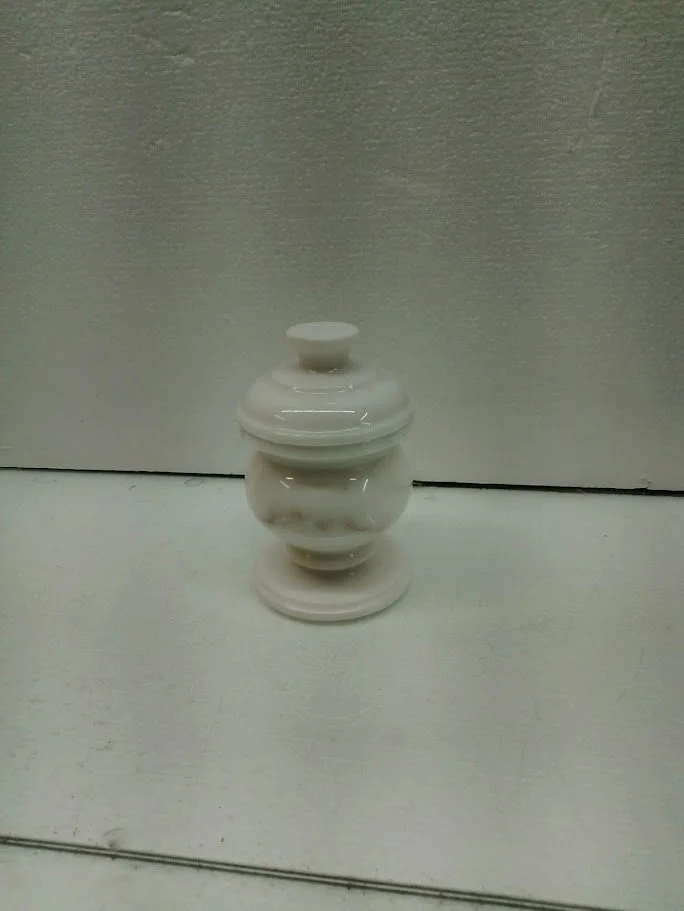 A white ceramic decorative jar with a lid, featuring a ornate, textured design, placed on a white surface against a plain, light-colored wall.