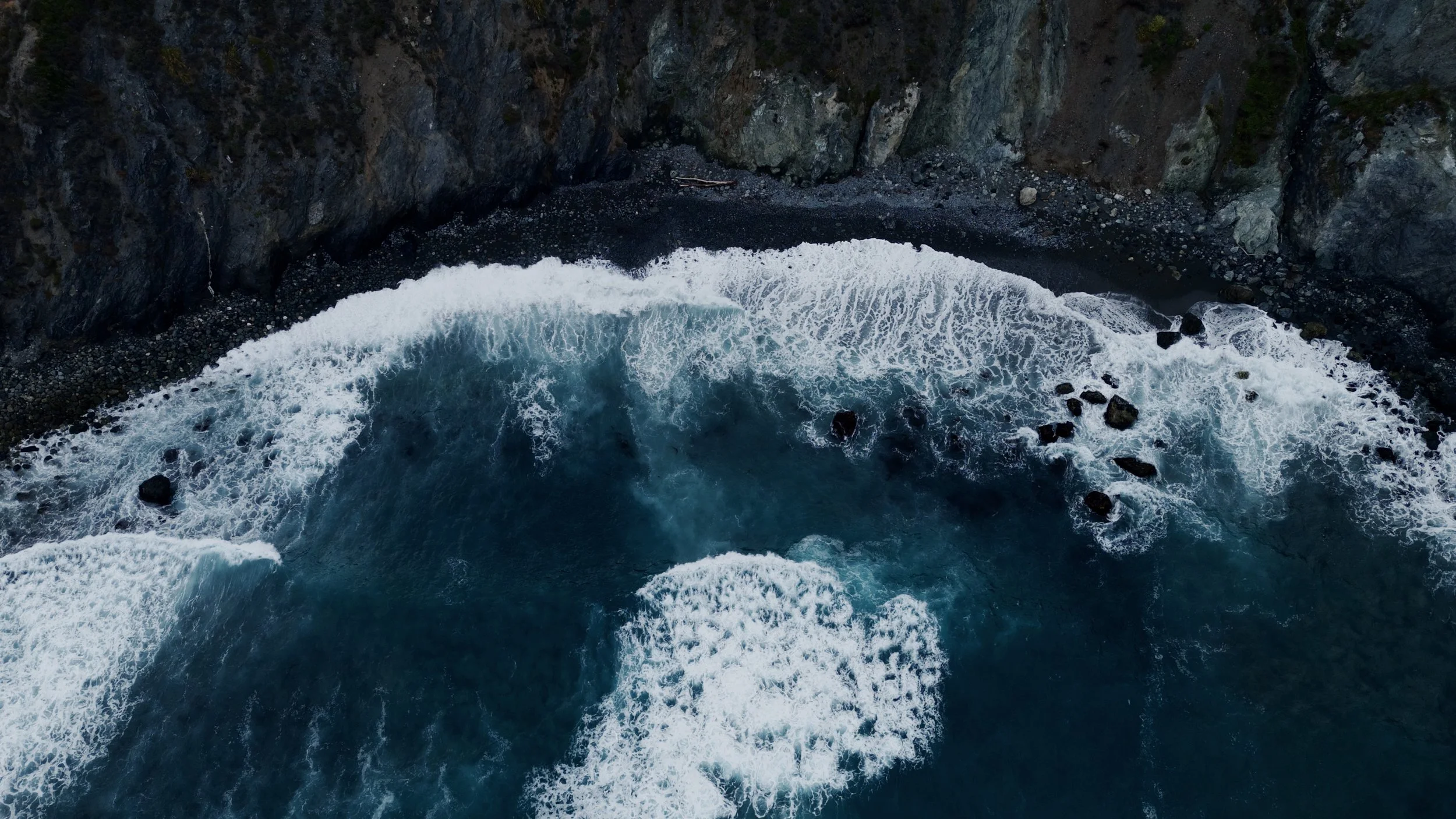 Aerial view of crashing ocean waves against a rocky shoreline with cliffs in Big Sur, California, USA.