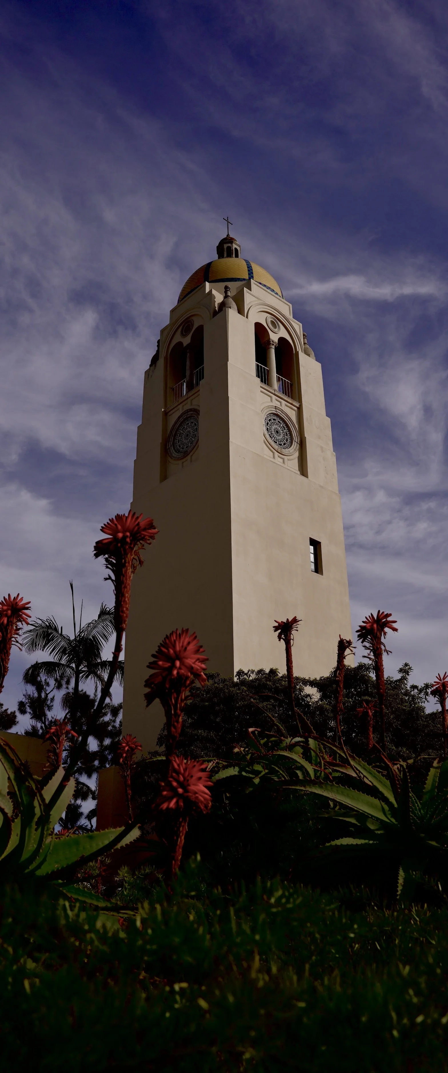 A tall clock tower with a yellow and blue dome on top, set against a partly cloudy sky, with red flowers and green plants at the base.