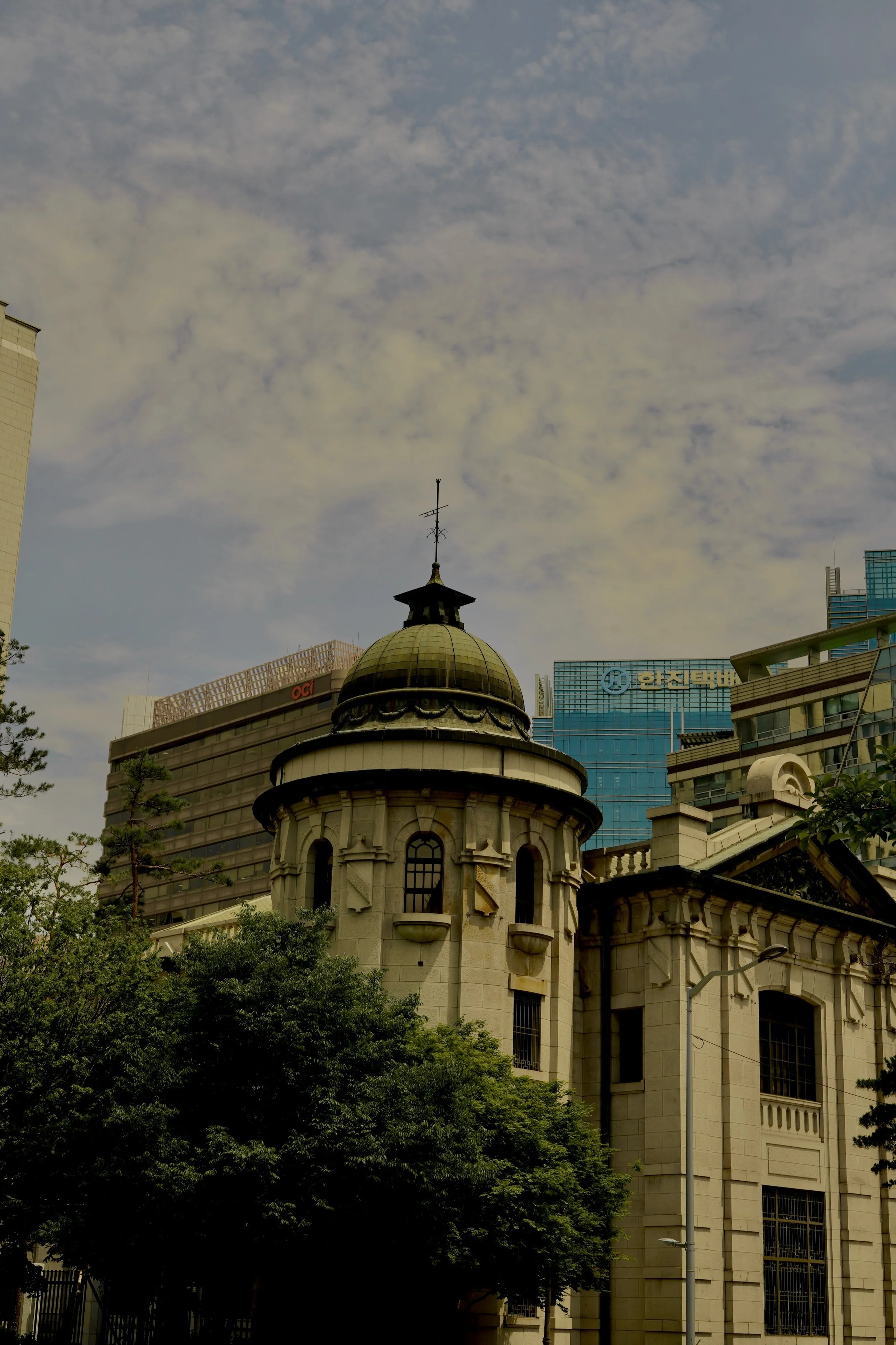 A historic building with a domed roof and a weather vane, surrounded by modern buildings and trees under a cloudy sky.