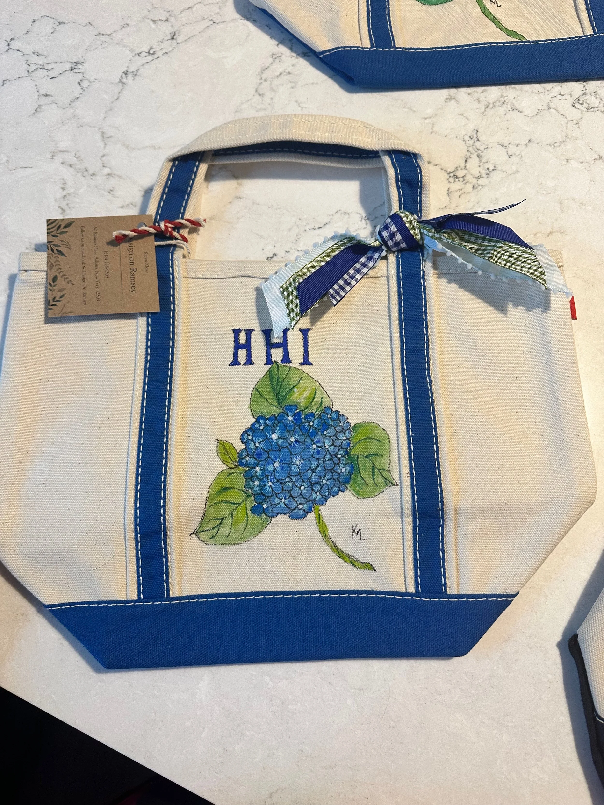 Cream-colored tote bag with blue accents, featuring a painted blue hydrangea flower, green leaves, and the word 'HI' embroidered above the flower, decorated with a blue and green checkered ribbon tied in a bow on the handle.