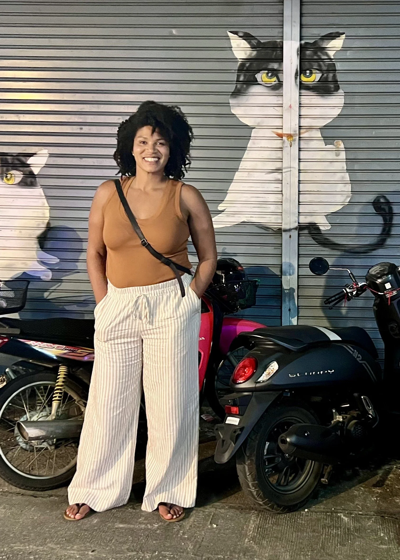 A woman with curly hair, wearing a tan tank top, white striped pants, and sandals, standing next to a pink scooter with a black color guard, in front of a metal shutter painted with large cat murals.