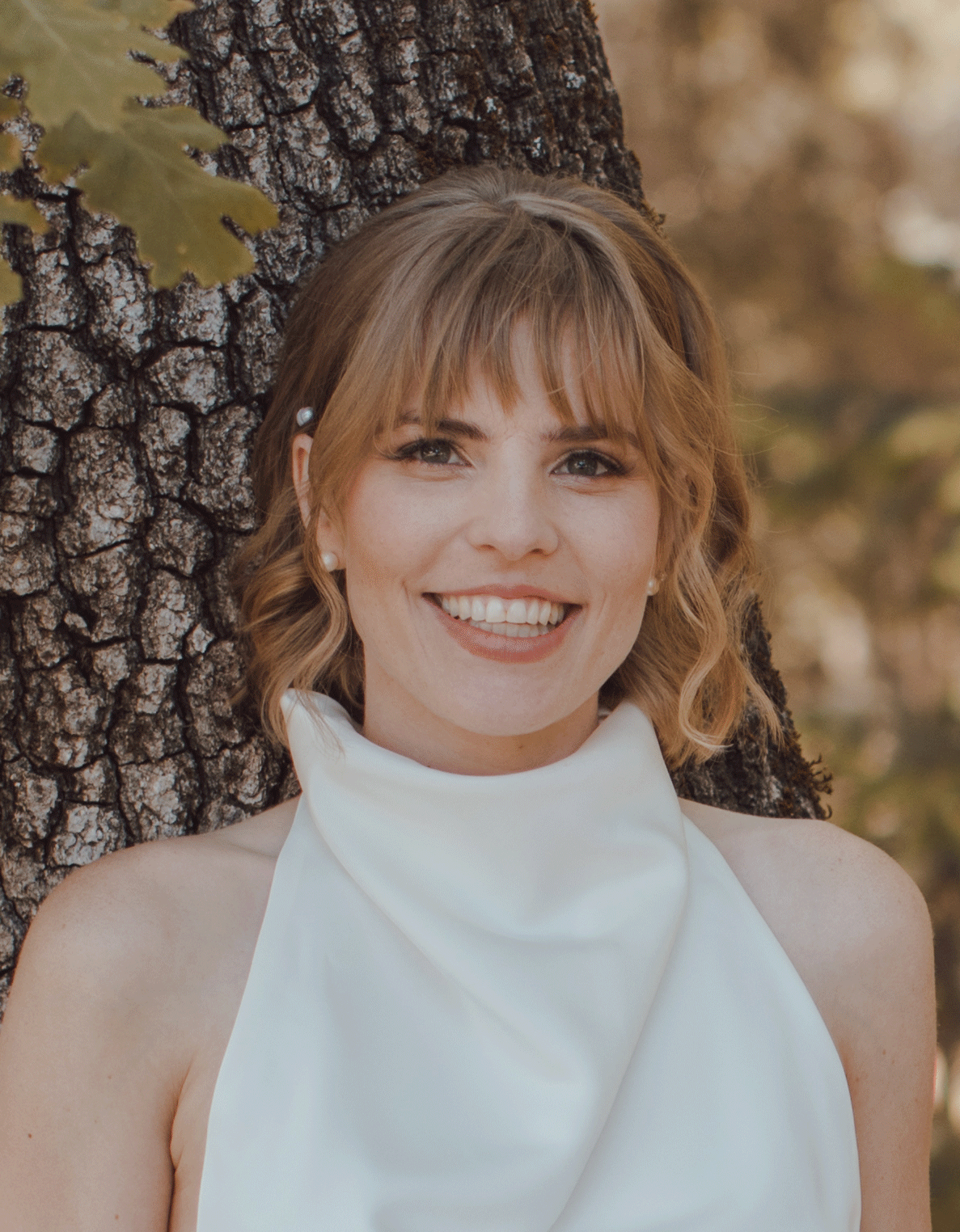 A woman with shoulder-length light brown hair and bangs, smiling, standing outdoors near a tree with dark textured bark, wearing a white sleeveless top with a high collar.