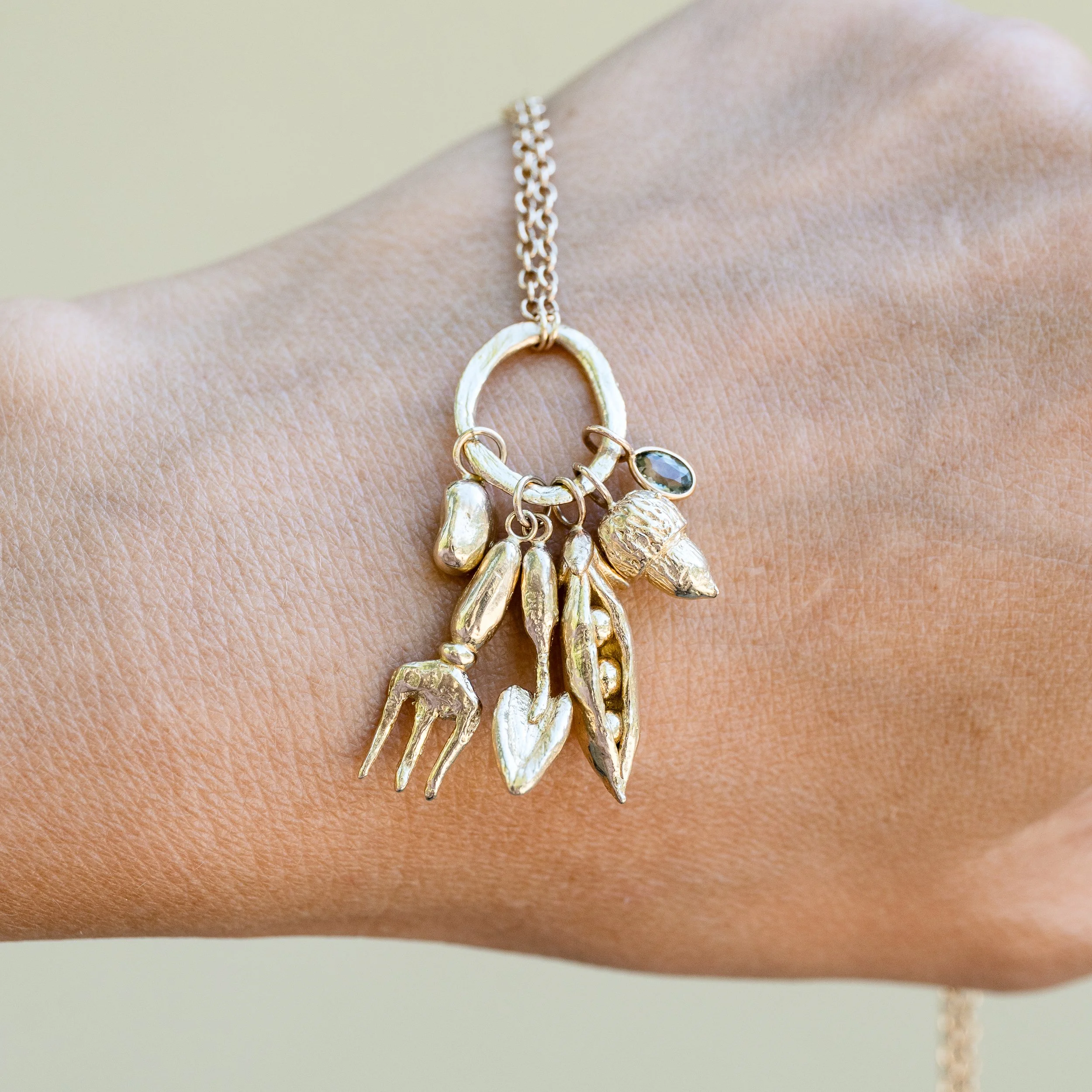 Close-up of a woman's wrist holding a gold necklace with charms, including an acorn, pea pod, fork and trowel and a bean, against a light-colored background.