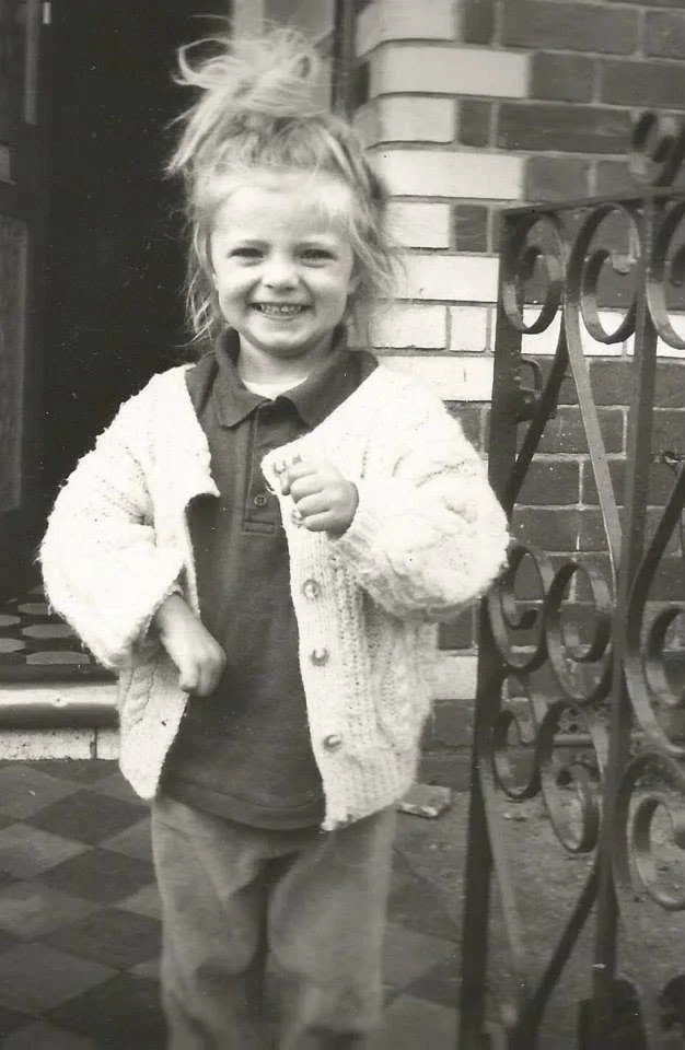 A black and white photo of a 5 year old girl with messy hair smiling at the camera, standing outside near a brick wall and a decorative iron gate.