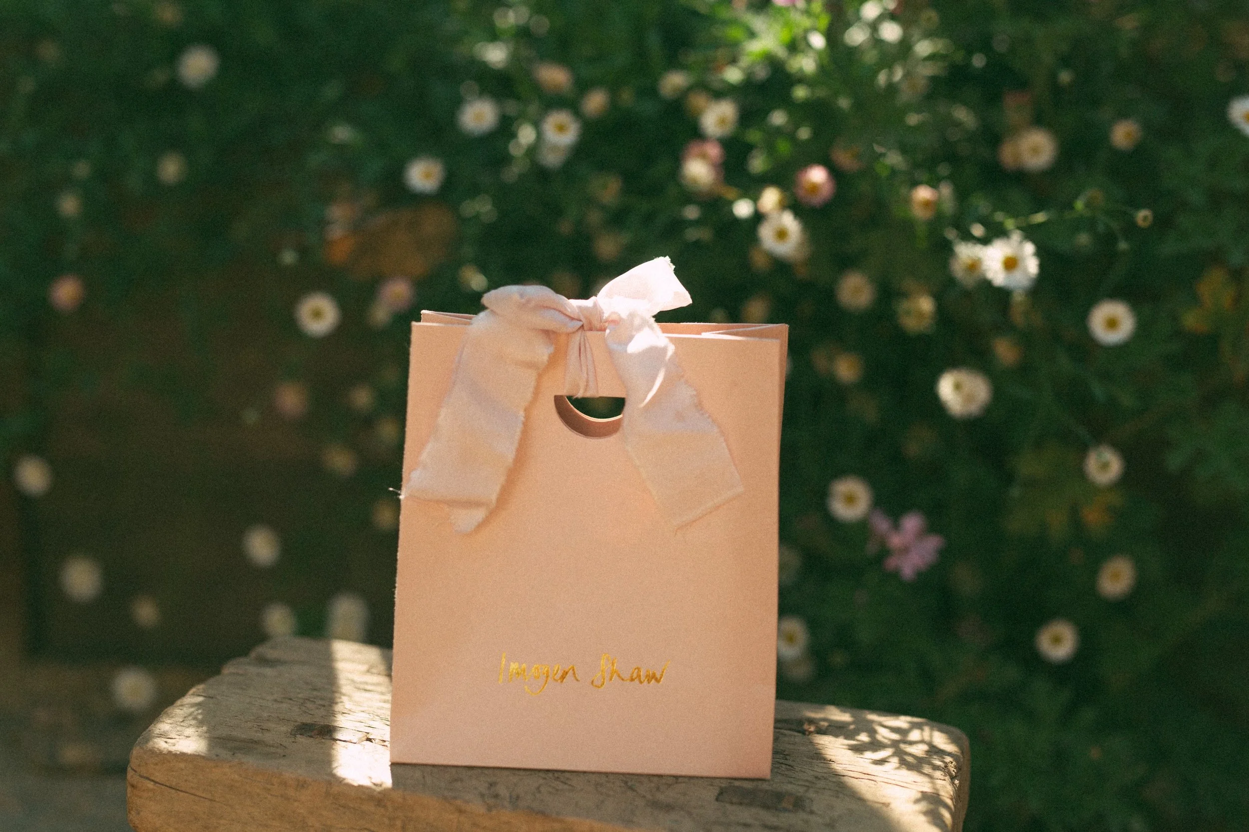 Pink paper jewelry bag tied with silk, 'Imogen Shaw' written in gold handwriting on the bag