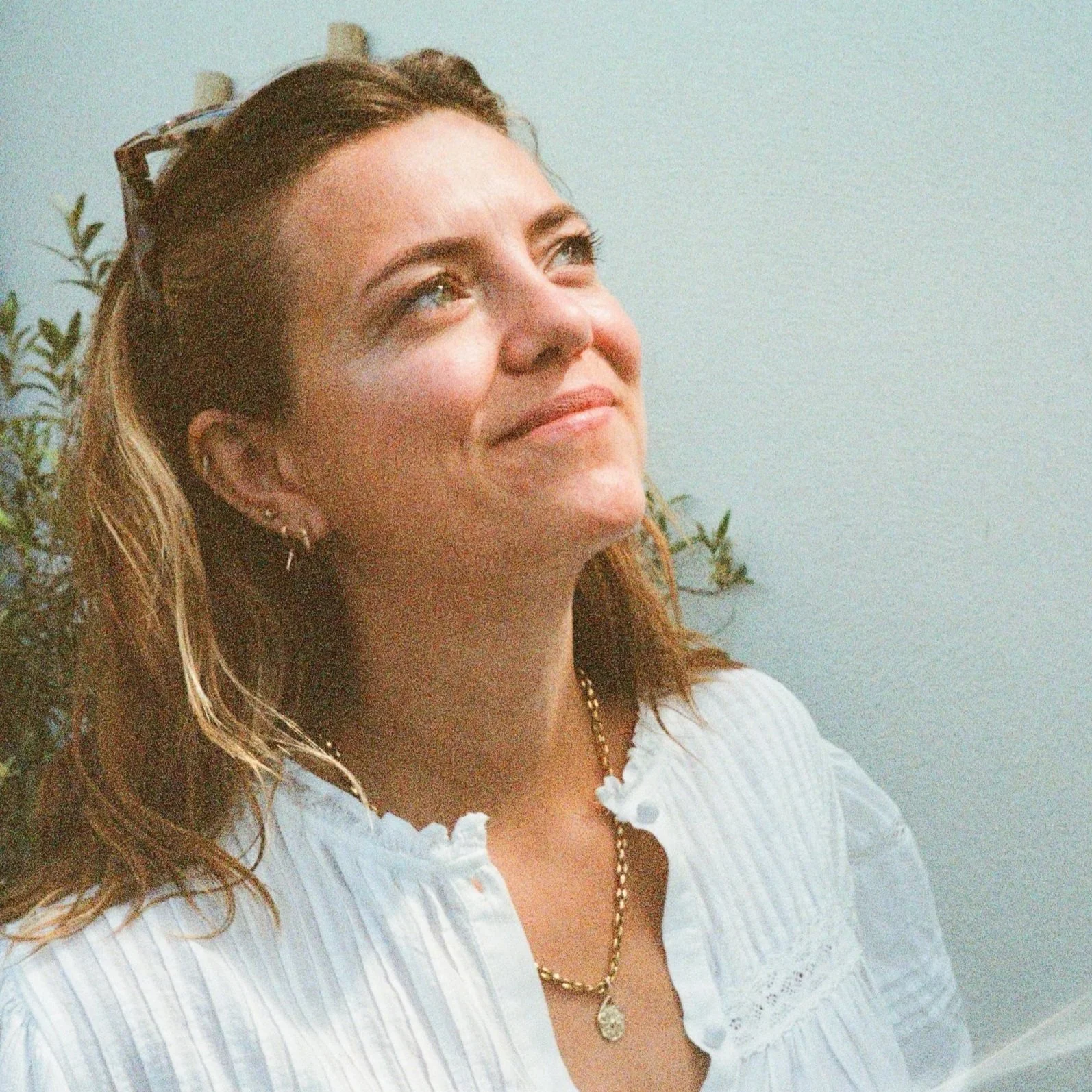 A woman with brown wavy hair, wearing a white top with a ruffled collar, sunglasses on her head, earrings, and a gold necklace with a pendant, smiling and looking up.