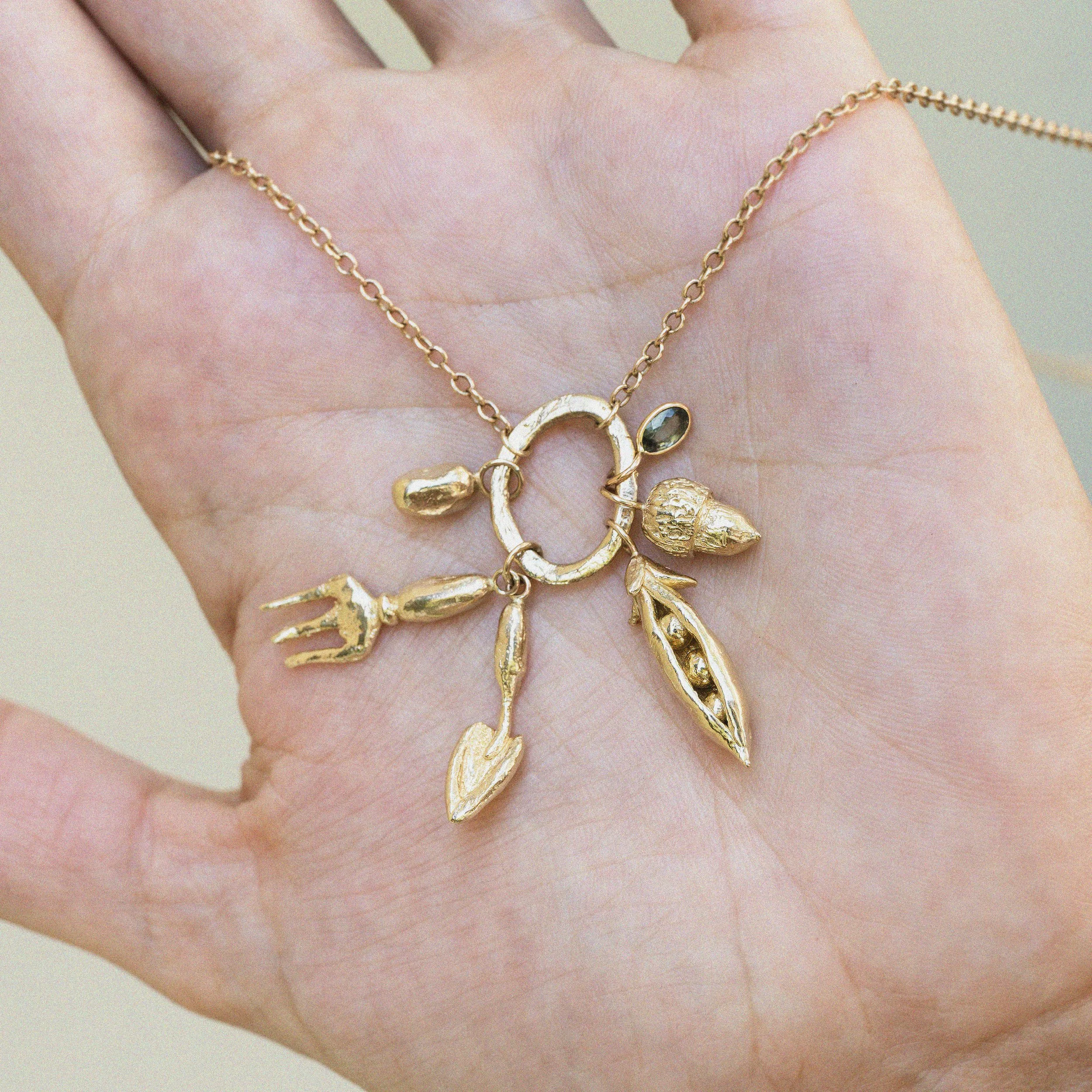 Close-up of a person's hand holidng a gold necklace with multiple charms, including an acorn, pea pod, fork and trowel and bean.
