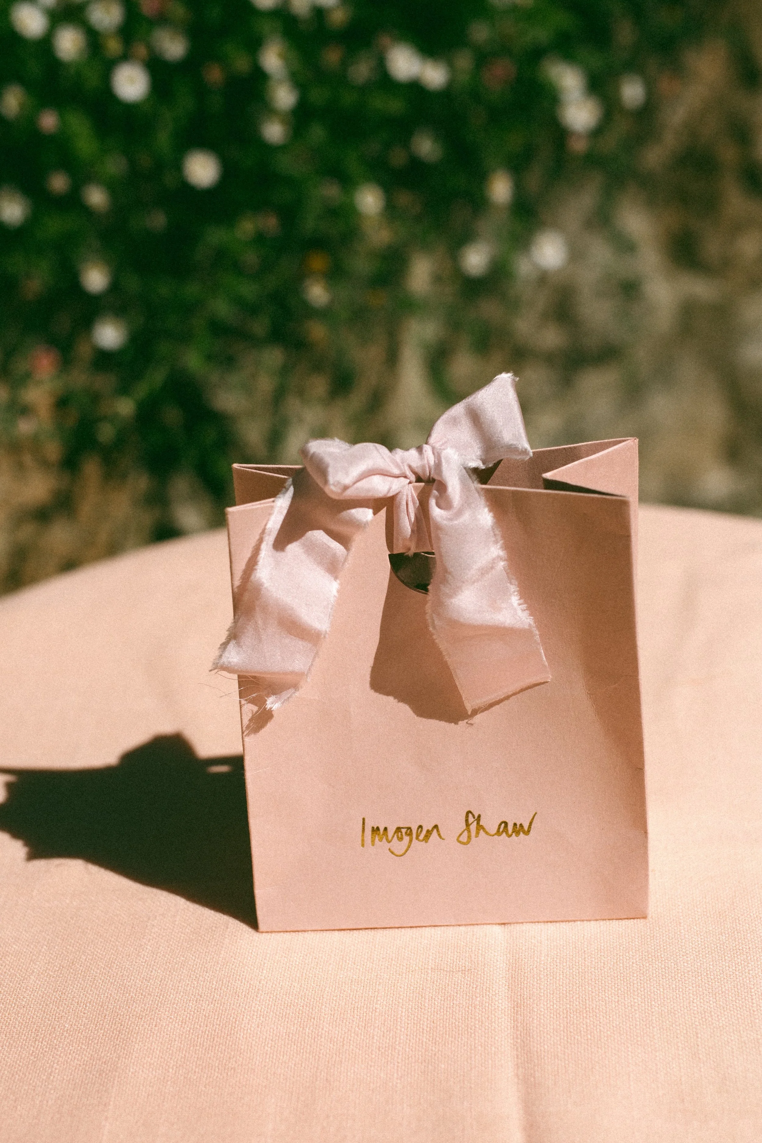 Pink paper jewelry bag in front of daisies, tied up with silk, Imogen's logo written in gold handwriting on the bag
