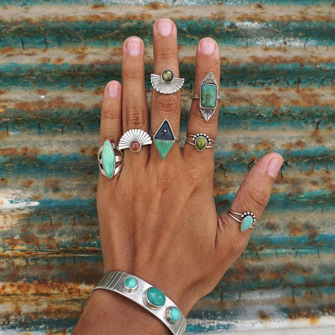 A variety of silver rings featuring green stones and tribal inspired detailing on a hand with a silver and turquoise bangle in front of a turquoise metal background