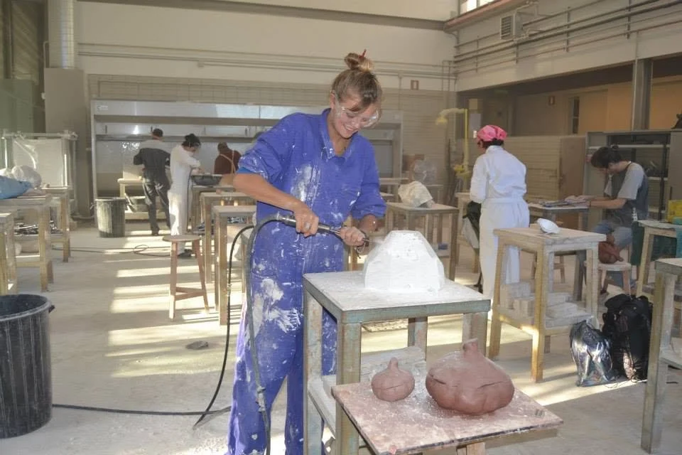 A woman in a blue jumpsuit is smiling and wearing safety glasses in a stone sculpture workshop with other people working on similar projects.