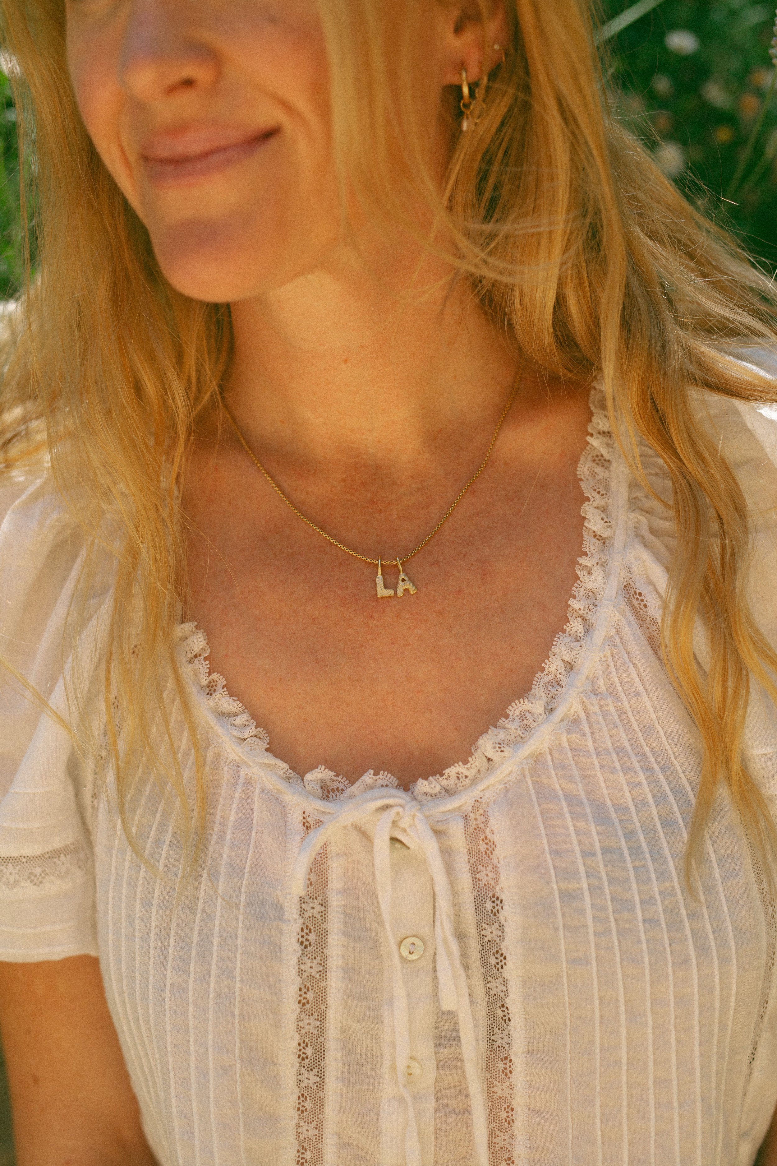 Close-up of a woman with long blonde hair wearing a white, lace-trimmed blouse and a necklace with the fingerprint charm initials 'LA' in gold, outdoors with green foliage in the background.