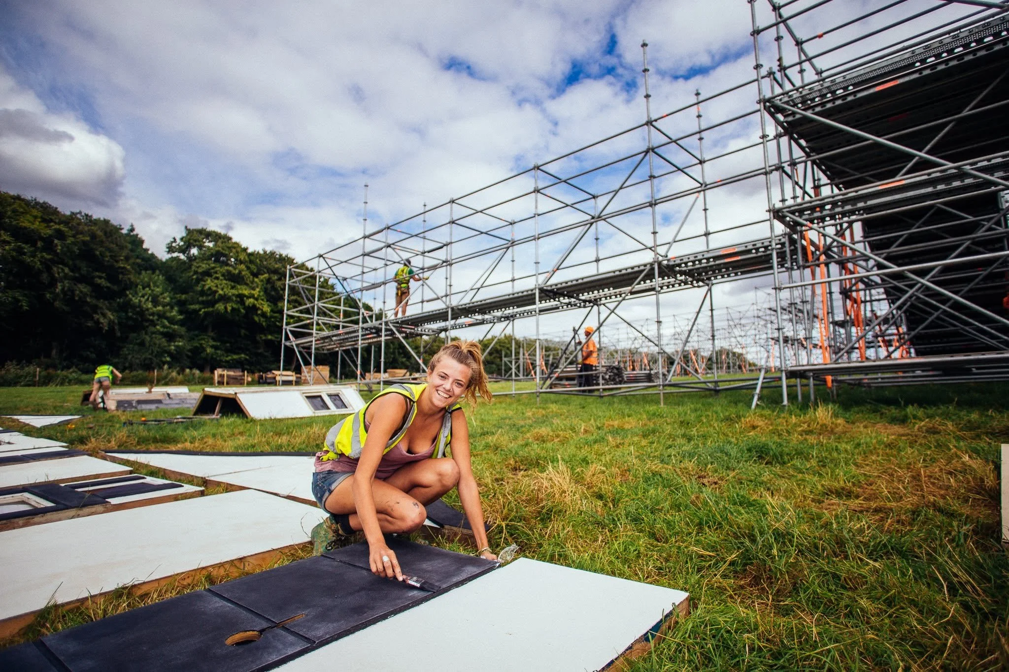 A woman with a yellow safety vest crouches on the ground painting a panel during construction work outdoors, with scaffolding and other workers in the background under partly cloudy sky.
