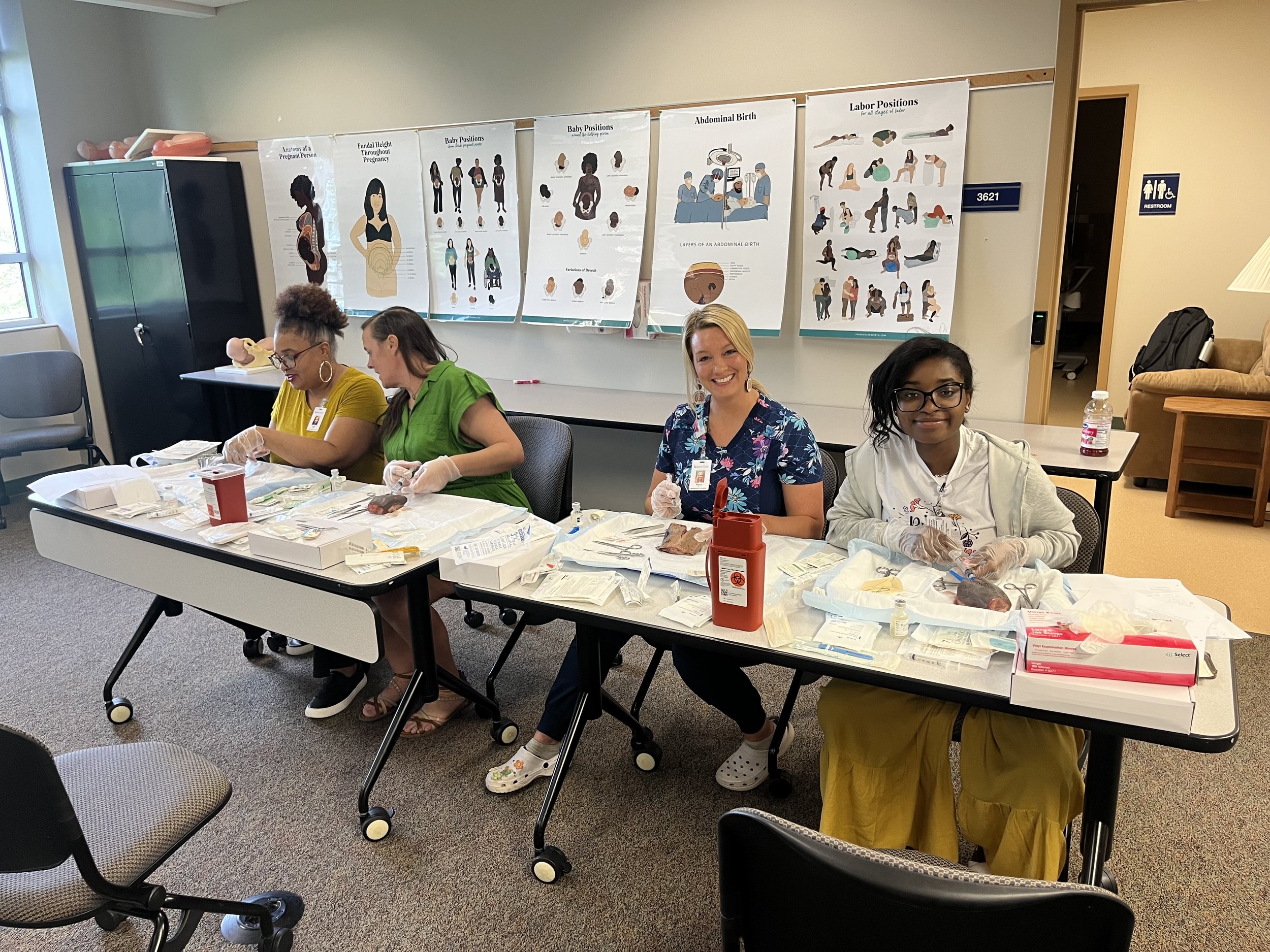 Four women sitting at a table during a childbirth education class or workshop, with anatomical posters of pregnancy, baby positions, and labor positions on the wall behind them.