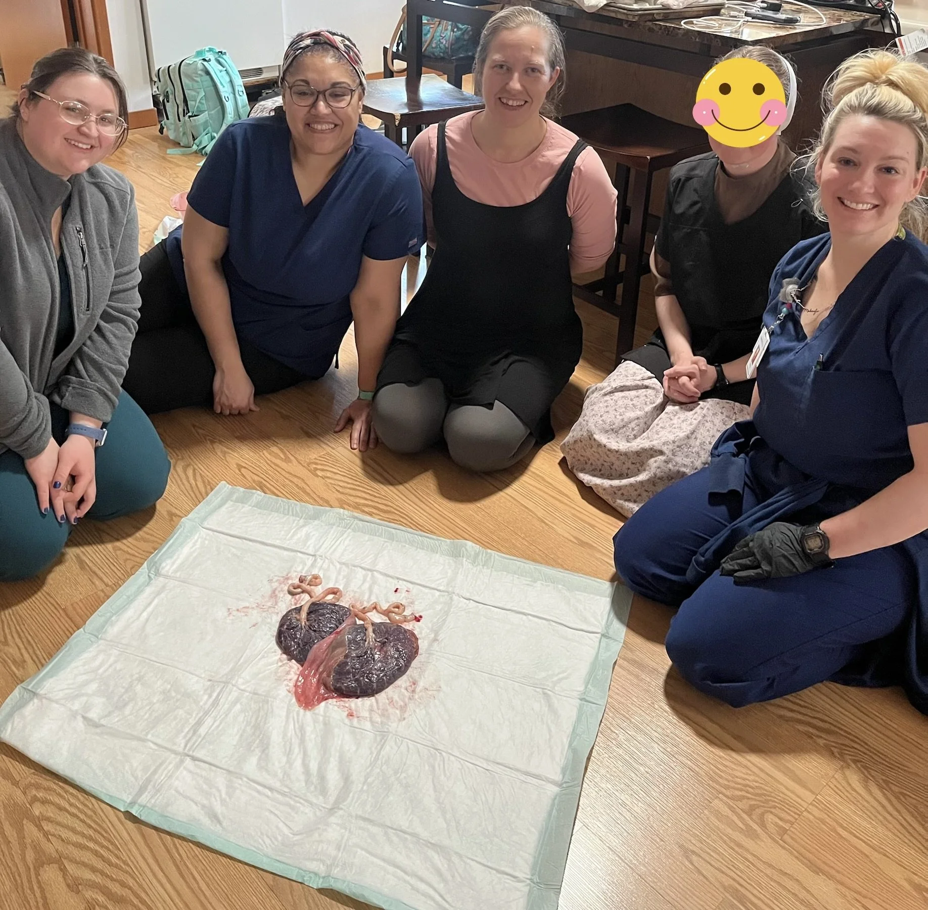 Group of six women sitting and kneeling on a wooden floor, smiling, around a mock placenta and fetus model on a white cloth. The women appear to be healthcare professionals, with some wearing scrubs or gloves, in a room with musical instruments in th