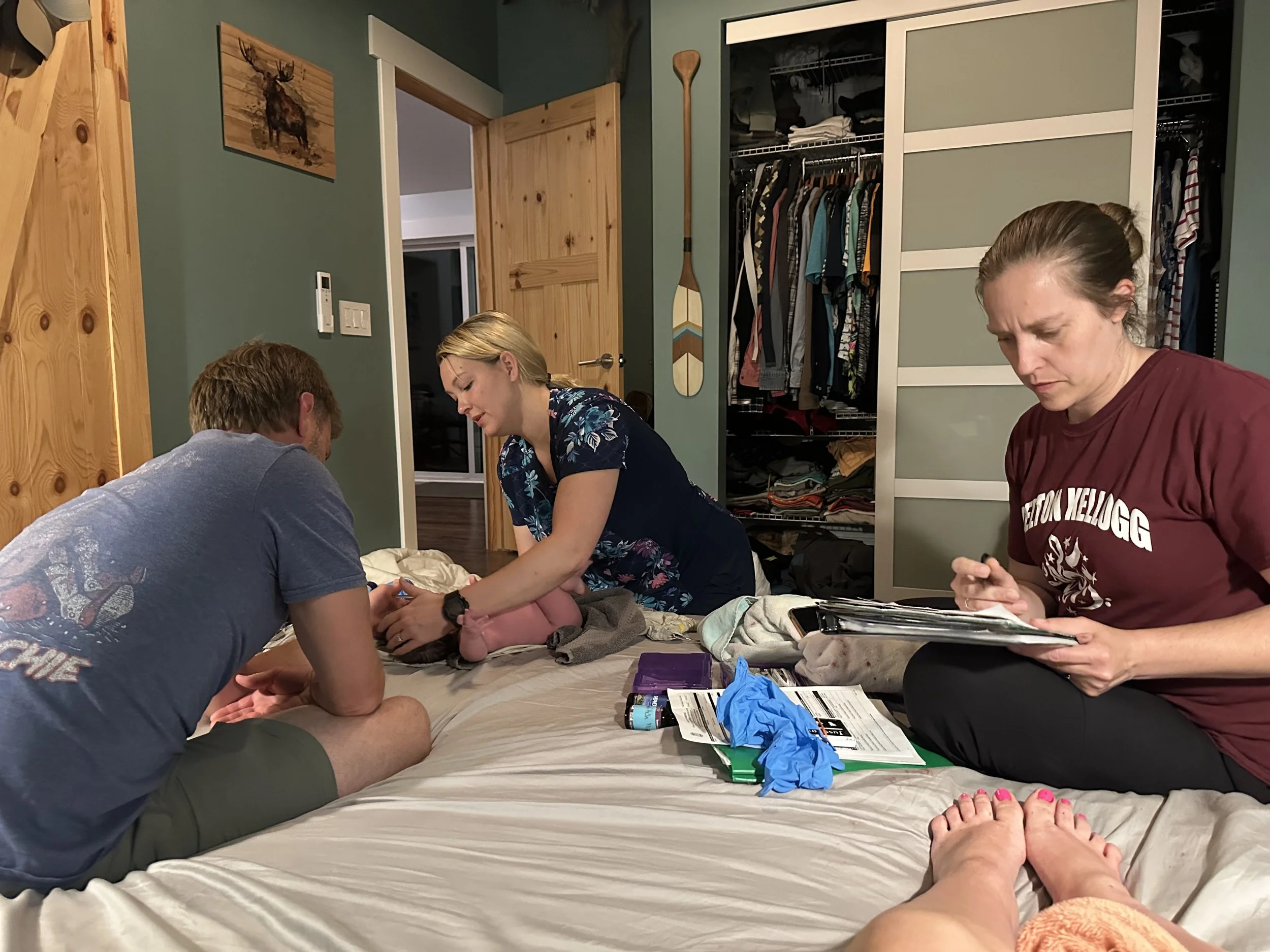A midwife woman attending to a newborn on a bed with two people sitting on either side, one taking notes and the other appearing to assist. The room has an open closet and wooden decor.