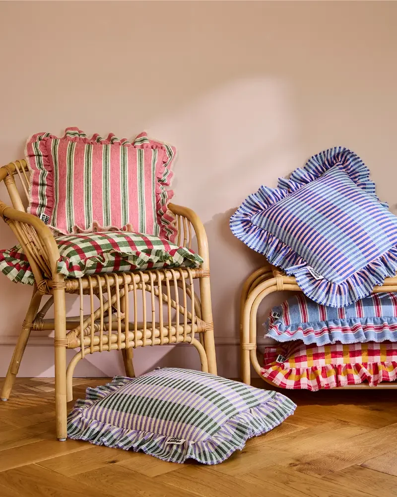 Rattan chairs with colorful striped and checkered cushions with ruffled edges, placed against a beige wall on a hardwood floor.