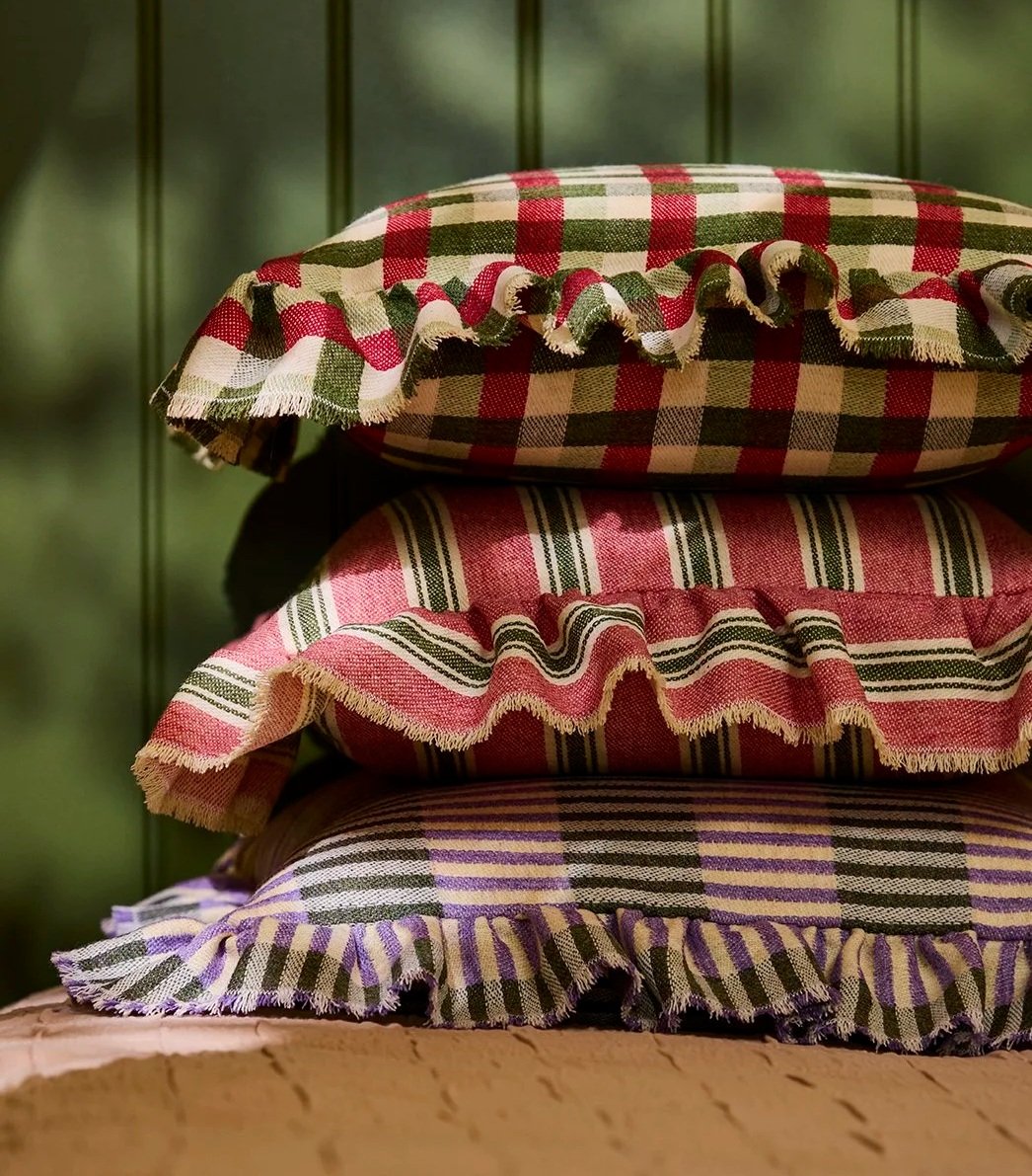 Stack of three ruffled cushions in green, pink, and purple on a bedspread in front of a panelled green wall.