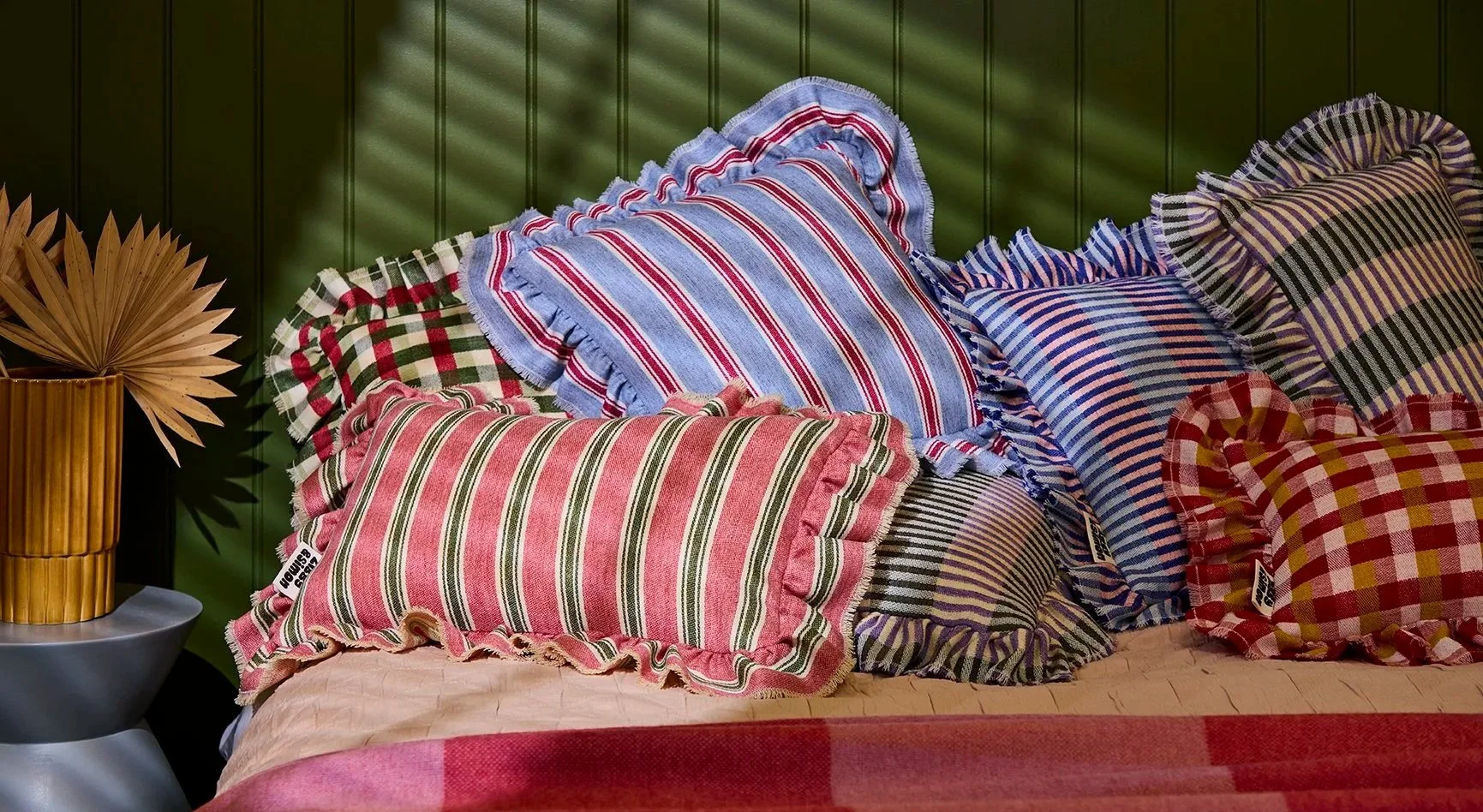 A bed with multiple striped and checkered pillows, a bedspread, set against a green-paneled wall. A decorative arrangement of dried palm leaves is on a side table to the left.