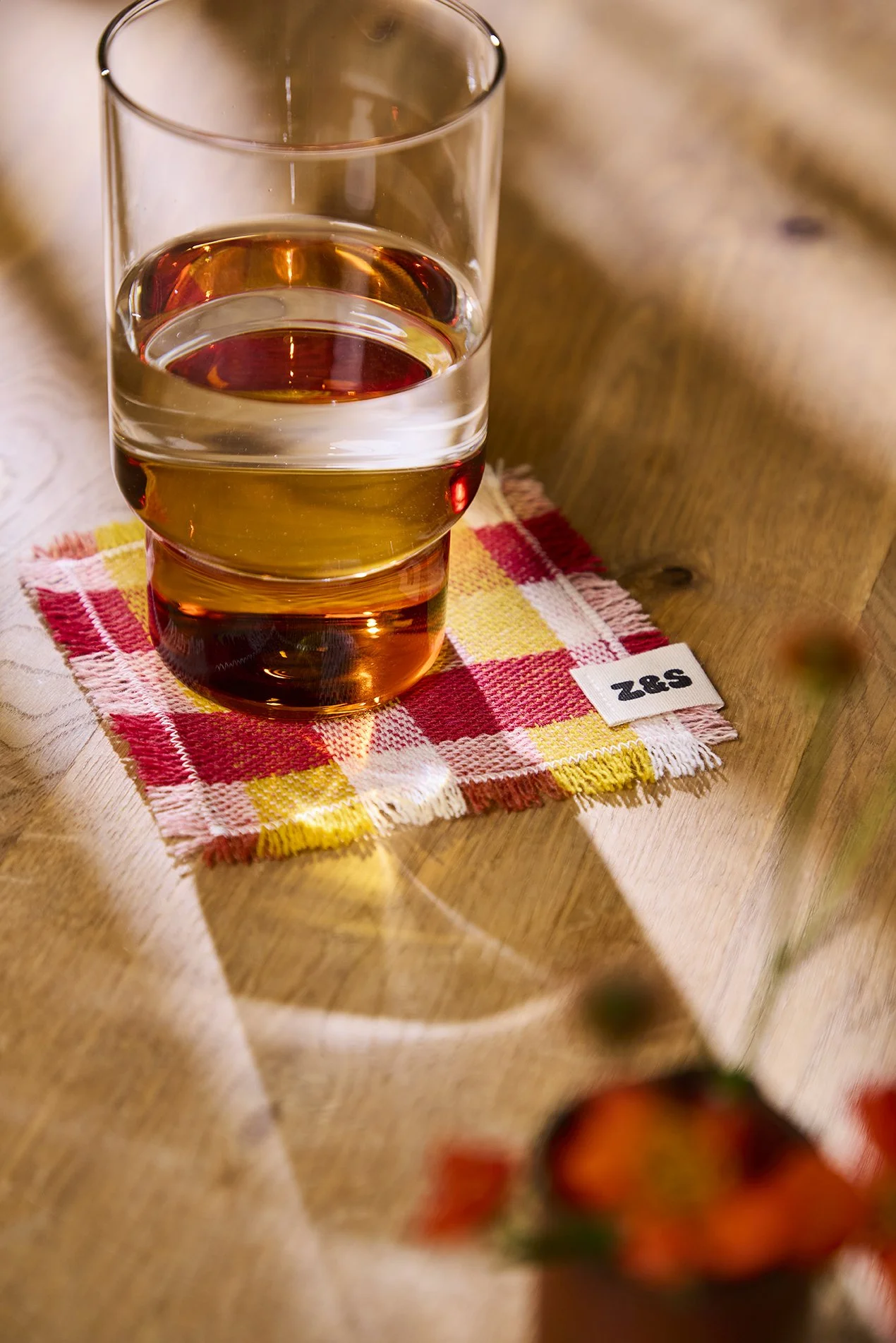 A glass of water on a red, yellow and white gingham coaster on a wooden table, with blurred red flowers in the foreground.