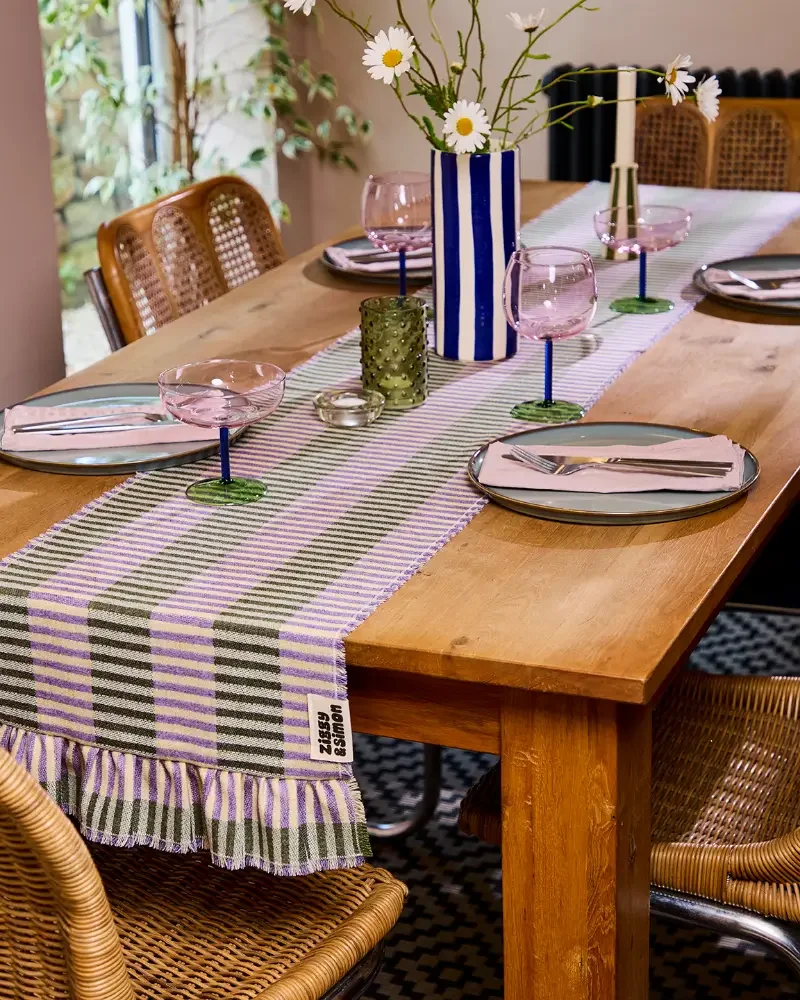 Dining table with a purple and green striped table runner, plates and wine glasses. The table has a wooden surface.