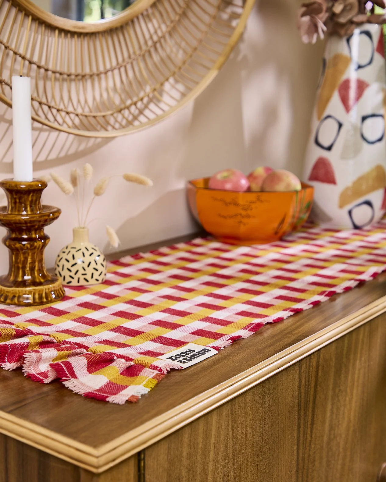 Decorative table with a red and yellow checkered table runner, a ceramic vase with dried flowers, a brown candlestick with a white candle, an orange bowl with apples, and a mirror above. The table has a wooden surface with a scalloped edge.