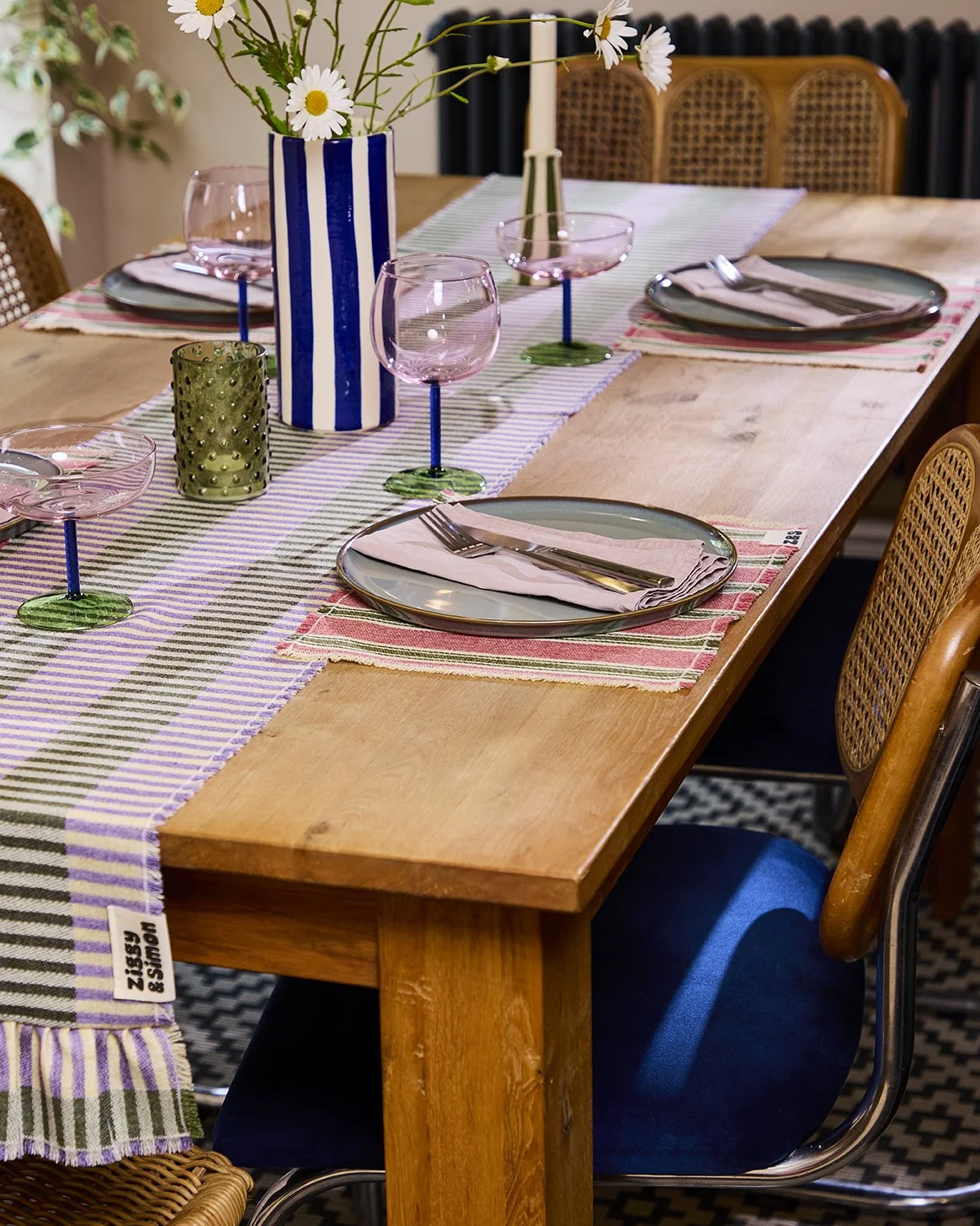 A wooden dining table set with place settings, including patterned placemats, plates, napkins, silverware, pink wine glasses, a striped blue and white vase with daisies, a tall white candle, and green candle holder. There are woven and upholstered chairs around the table.