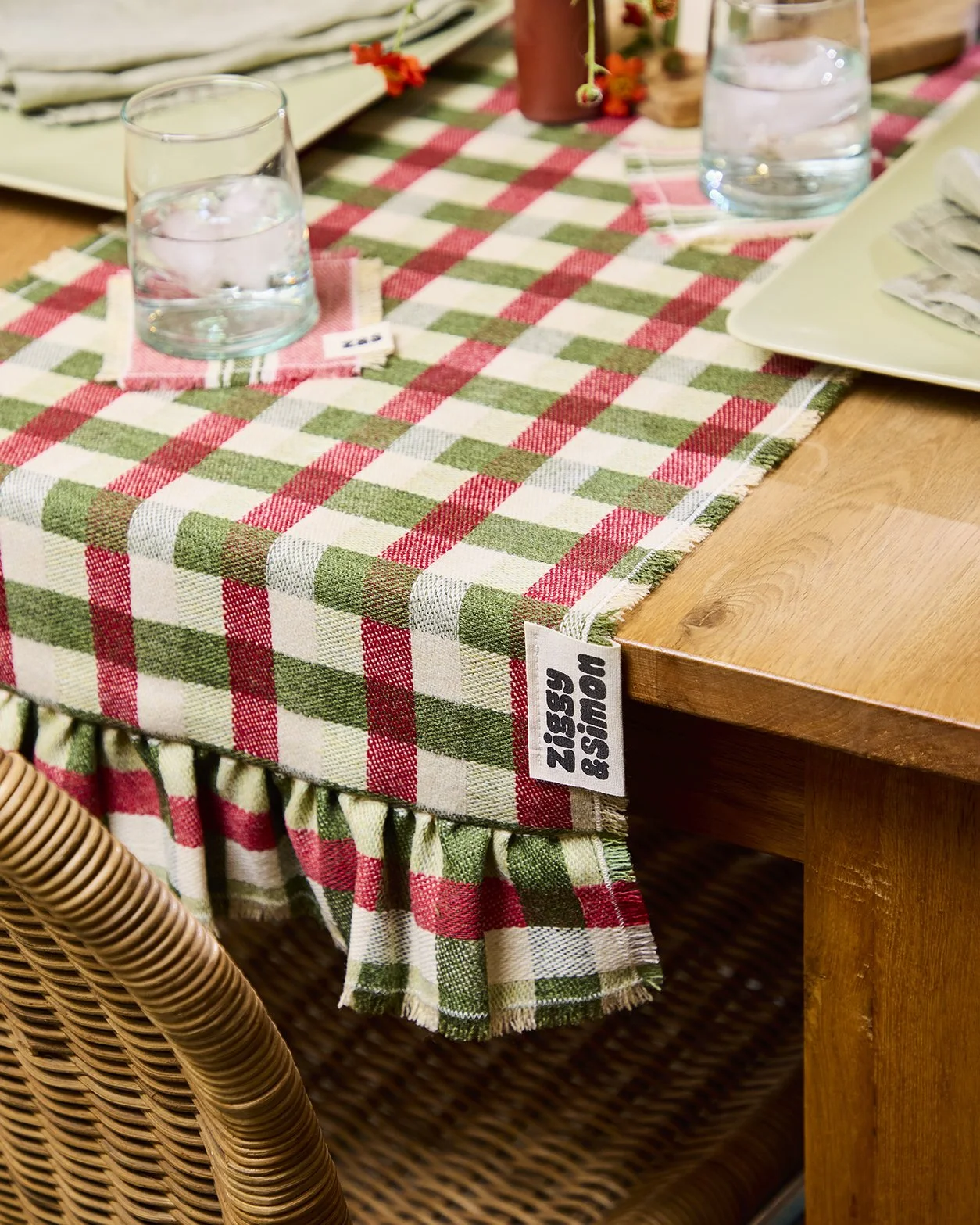 Checkered table runner with red, green, and white gingham pattern draped over a wooden table, with two glasses of water, plates, and a wicker chair partially visible.