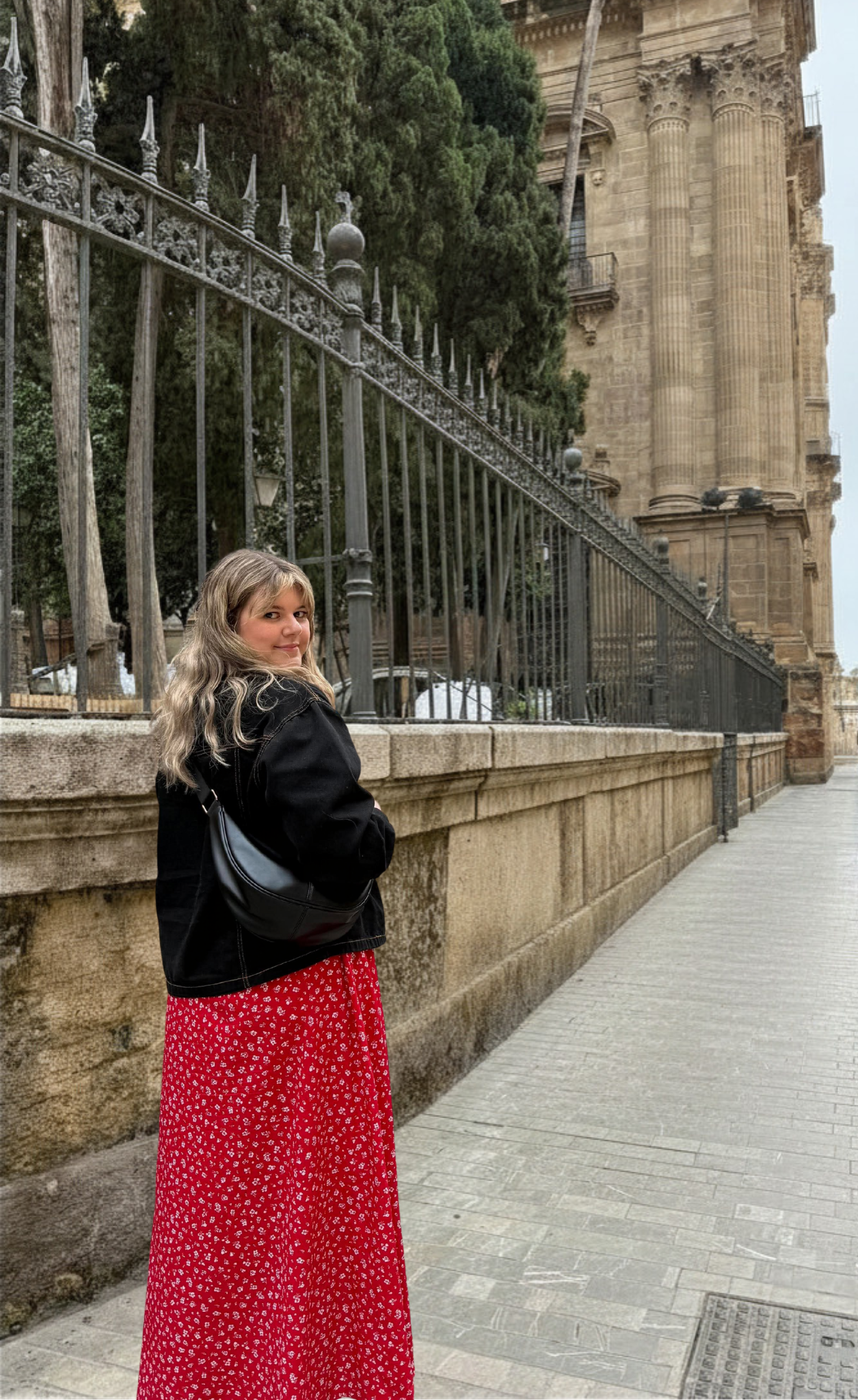 A woman with long blonde hair, wearing a red dress with white patterns, black jacket, and carrying a black handbag, standing on a sidewalk next to a stone wall and iron fence, with historical architecture and large trees in the background.