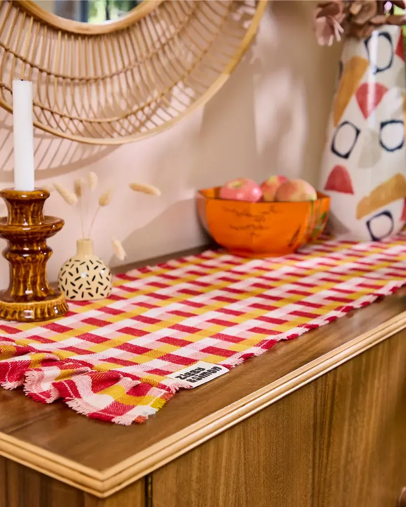 Decorative table with a red and yellow checkered table runner, a ceramic vase with dried flowers, a brown candlestick with a white candle, an orange bowl with apples, and a mirror above. The table has a wooden surface with a ruffled edge.