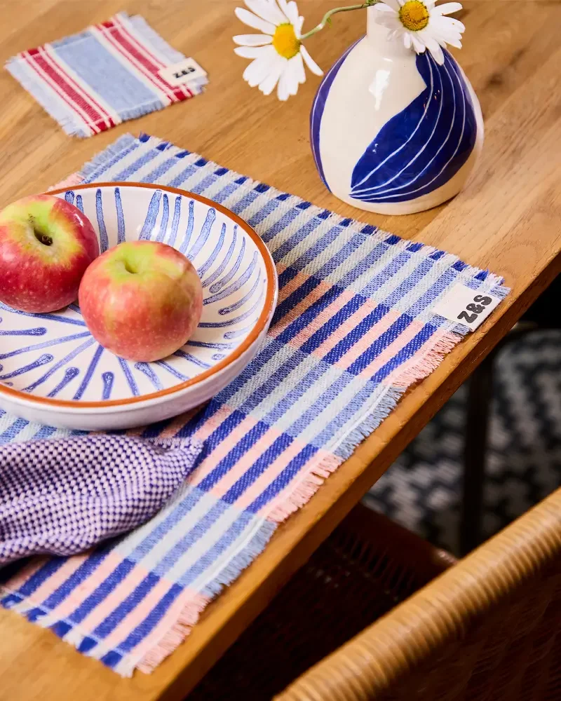 Two apples on a decorative plate, a ceramic vase with a blue and white abstract pattern holding a daisy flower, a pink, white, and blue striped placemat, and a striped coaster on a wooden table.