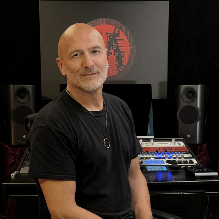 A man with a bald head, gray beard, and earrings, wearing a black t-shirt, standing in front of a music production setup with speakers, a laptop, and digital equipment, with a red and black digital artwork on a screen behind him.