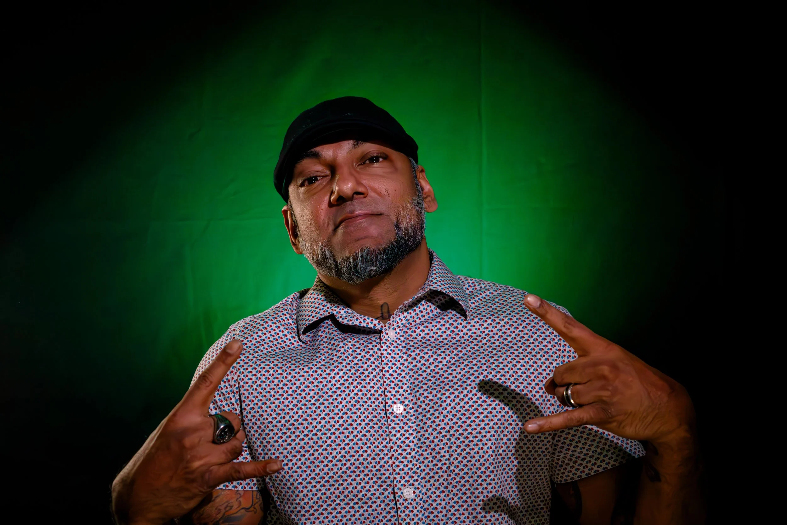 A man with a beard wearing a black cap and a patterned shirt, posing with both hands making wolf signs against a green and black gradient background.