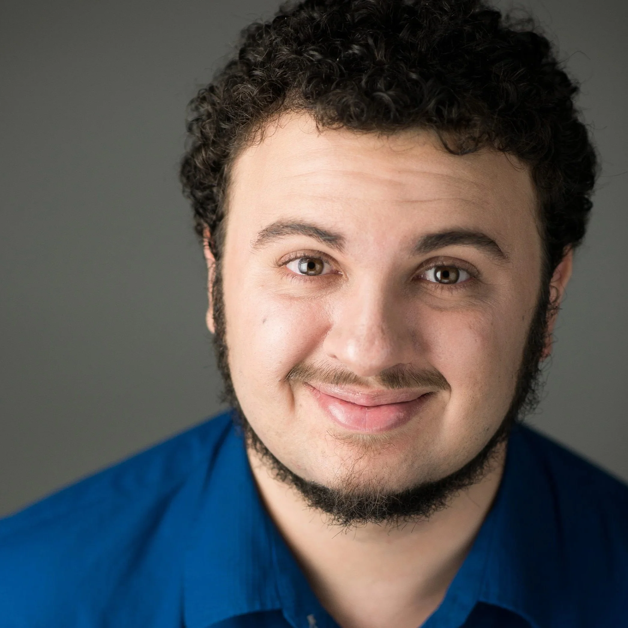Close-up portrait of a smiling man with curly dark hair, light skin, blue eyes, and a beard, wearing a blue shirt against a gray background.
