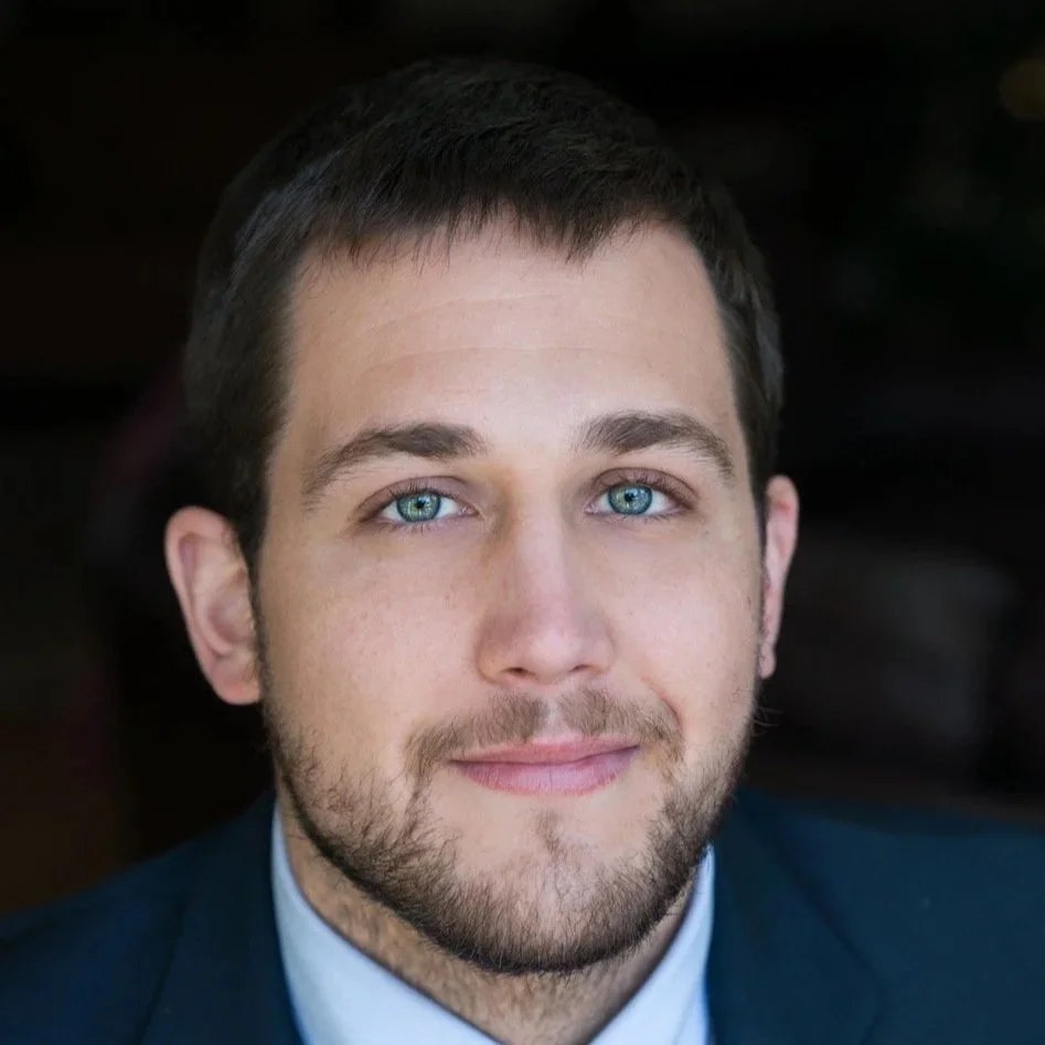 Close-up portrait of a young man with short dark hair, blue eyes, light skin, and a beard, wearing a dark suit and light blue shirt, smiling softly at the camera.