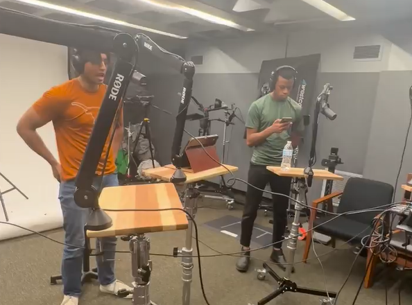 Two people in a recording studio, one wearing an orange shirt and the other in a green shirt with headphones, standing near microphones and audio equipment.