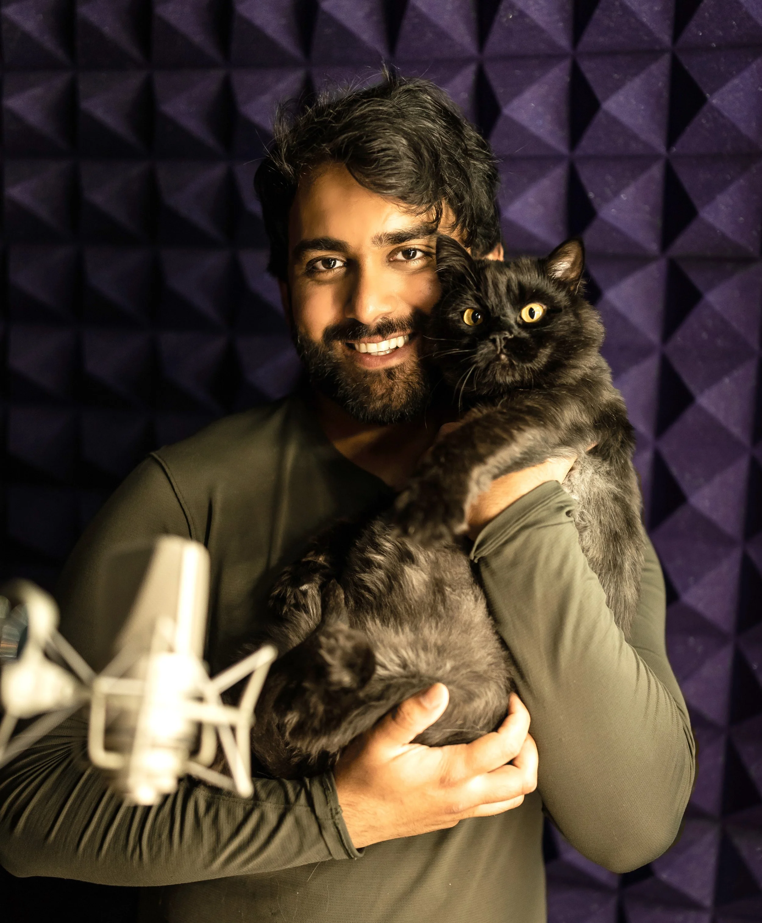 A man with dark hair and a beard smiling while holding a black cat in a recording studio with purple acoustic foam panels in the background.
