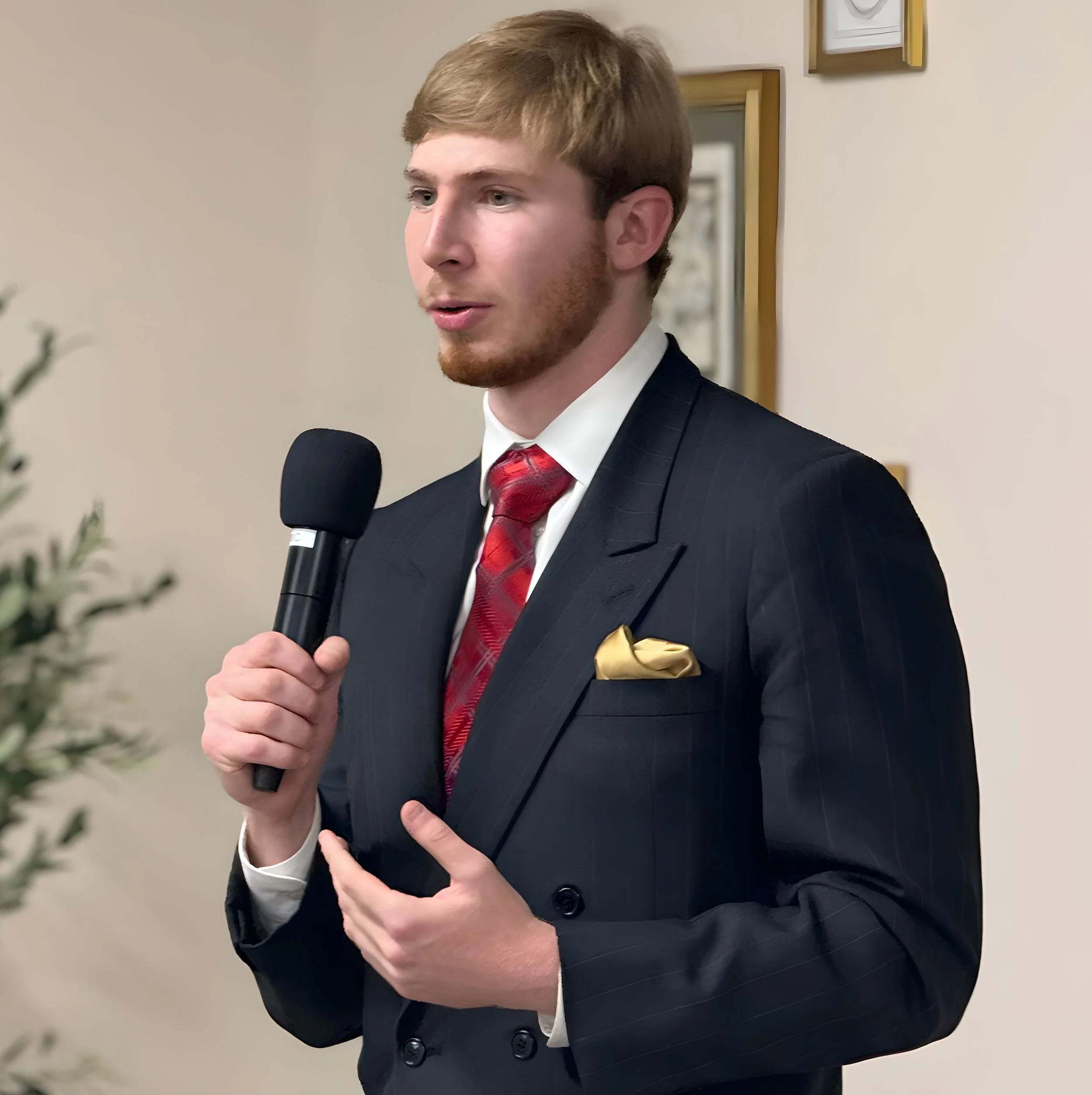 A young man in a dark business suit, white shirt, and red tie, holding a microphone, appears to be giving a speech or presentation in an indoor setting.
