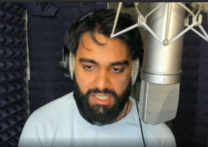 A man with dark hair and a beard wearing headphones, singing or speaking into a professional microphone in a recording studio with acoustic foam panels.