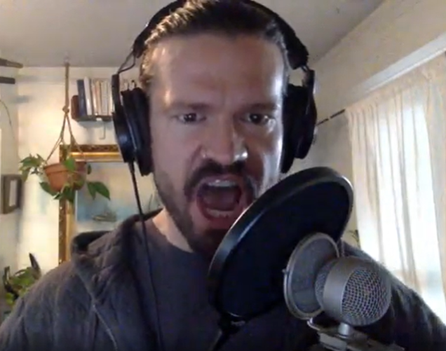 A man with a beard and dark hair wearing headphones, speaking or singing into a microphone in a home setting.
