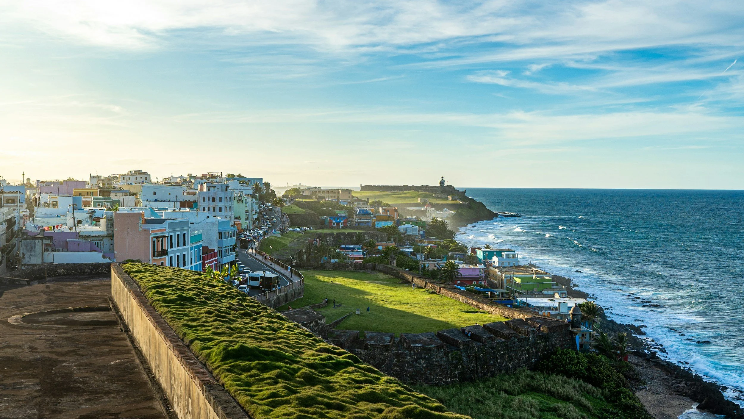 Colorful houses along a coastal street with a grassy area, a lighthouse in the distance, and the ocean on the right side.
