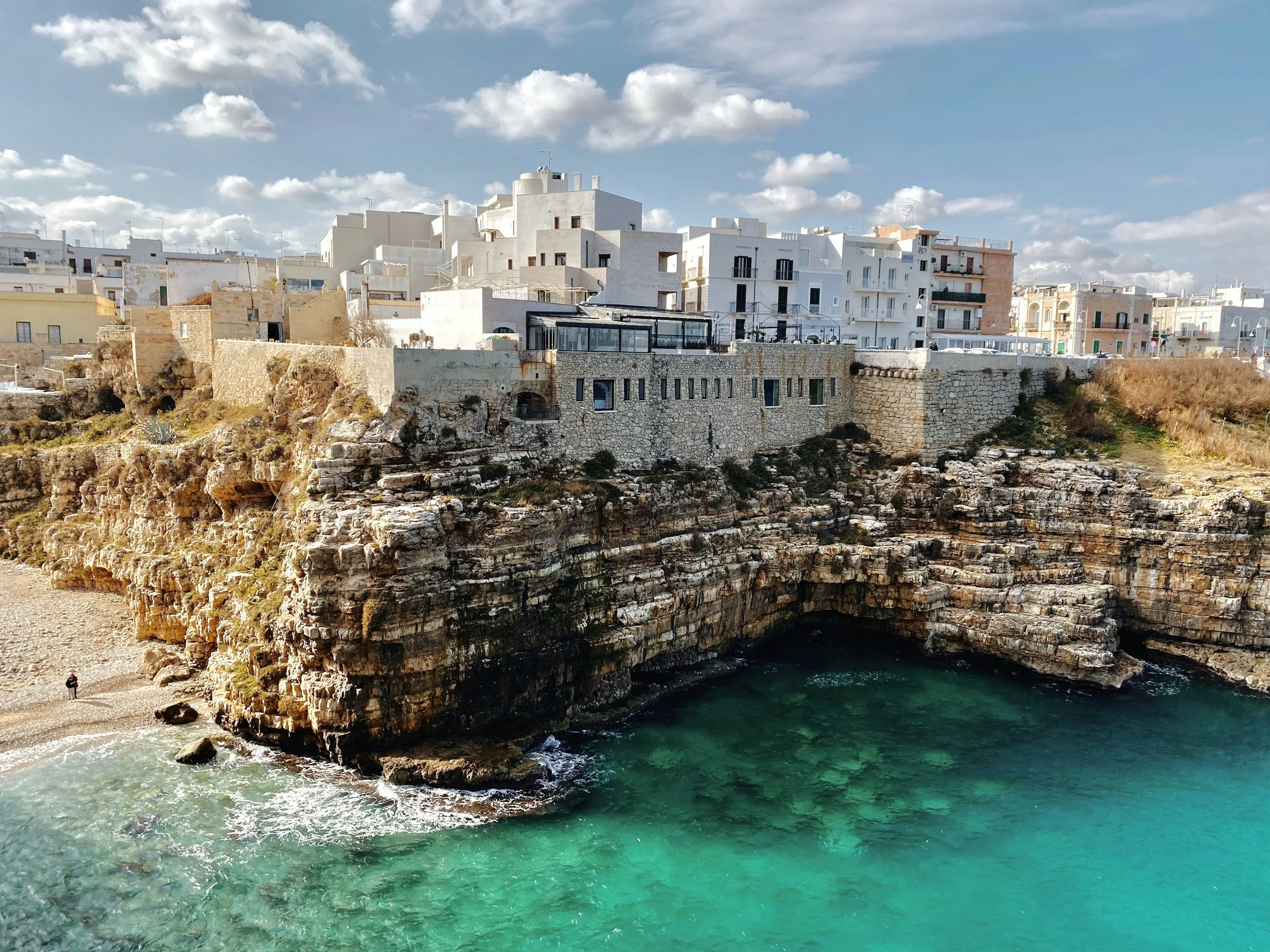 Cliffside with white buildings overlooking a turquoise ocean and a rocky cove, under a partly cloudy sky.