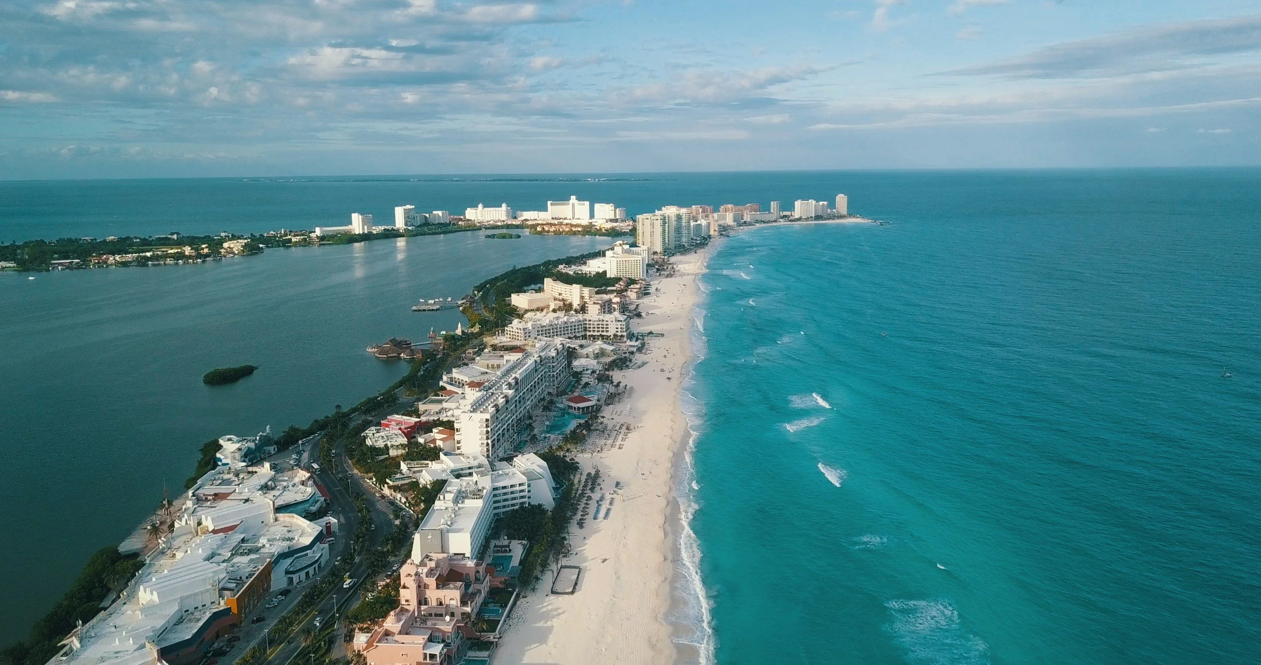 Aerial view of a beachside city with white sandy beach, turquoise ocean on the right, and a river or lagoon on the left, with numerous buildings and high-rises along the coast.