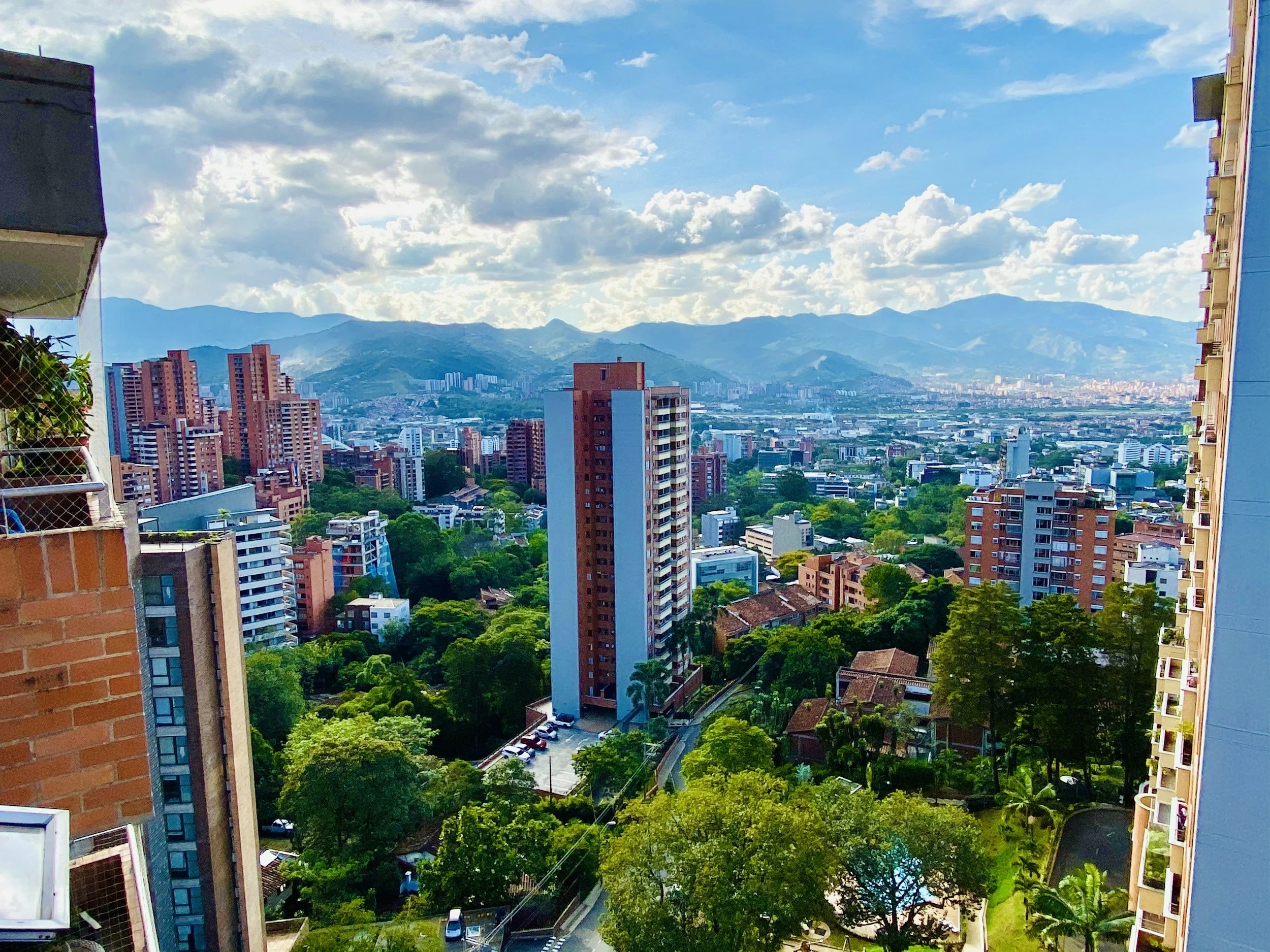 City skyline with tall buildings, lush green trees, and mountains in the background under a partly cloudy sky.