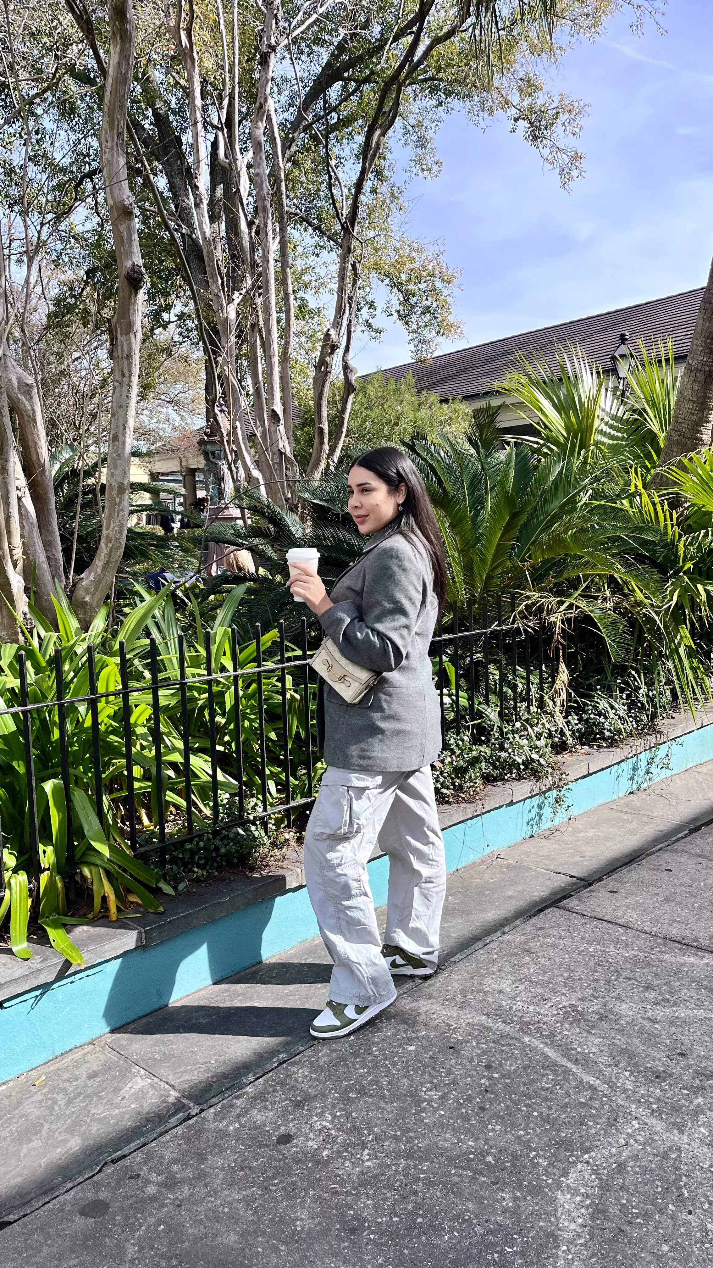 A young woman with dark hair wearing a gray blazer, white cargo pants, and sneakers, holding a disposable coffee cup, standing beside a low fence with lush green plants and trees in the background on a sunny day.