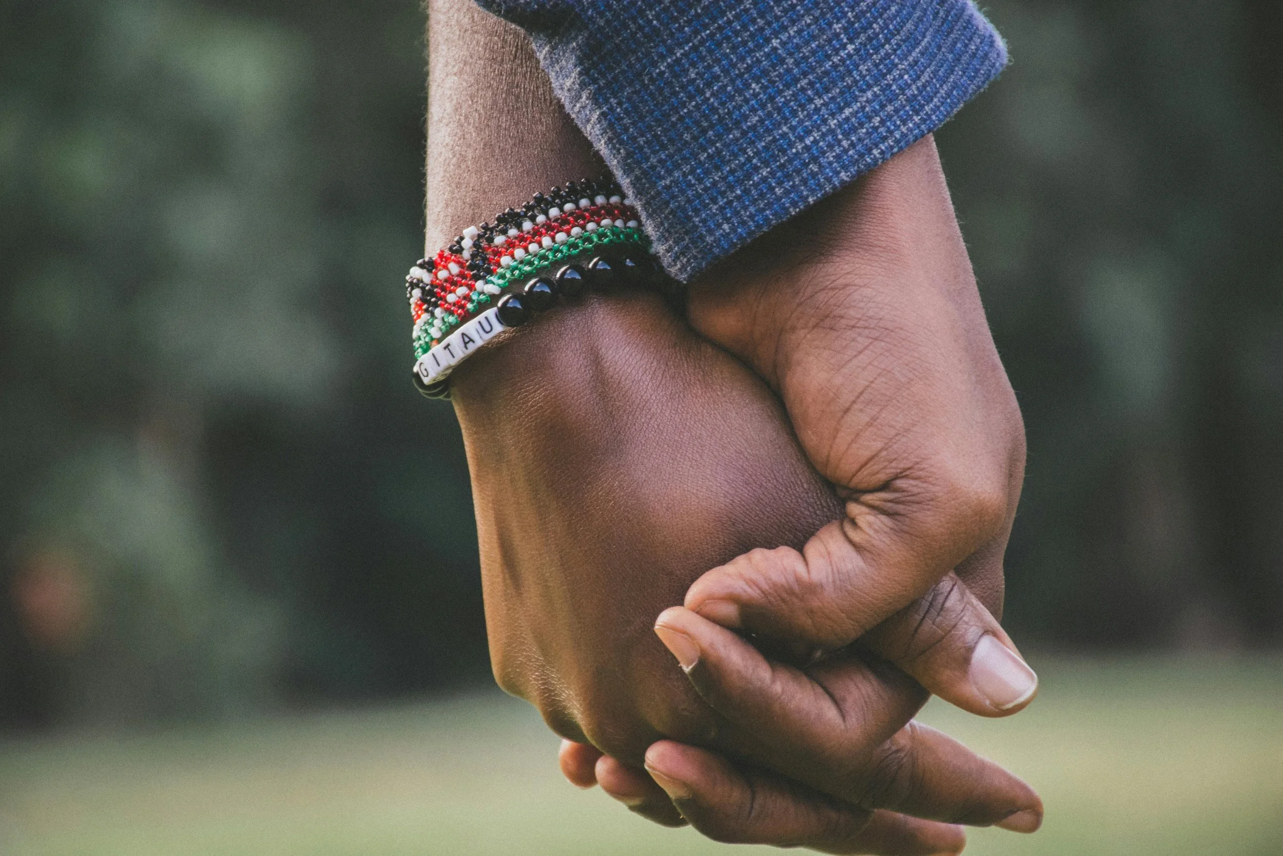 Close-up of two people holding hands outdoors, one person wearing multiple colorful beaded bracelets, and the other person’s hand partially visible.