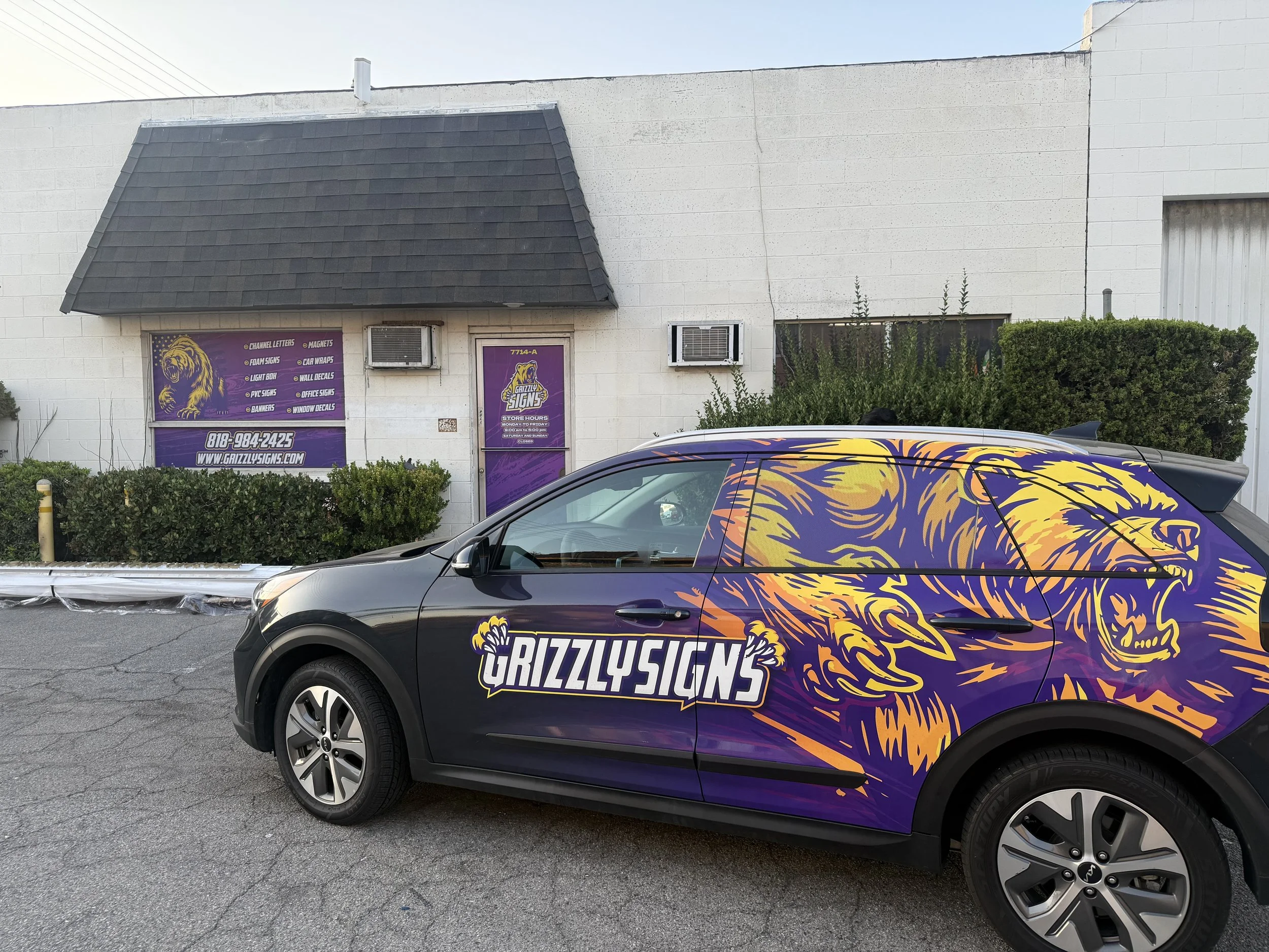 Black vehicle with a colorful lion graphic and the words 'Grizzly Signs' parked in front of a building with purple signage and bushes. | Grizzly Signs | Los Angeles, CA | Custom Wraps