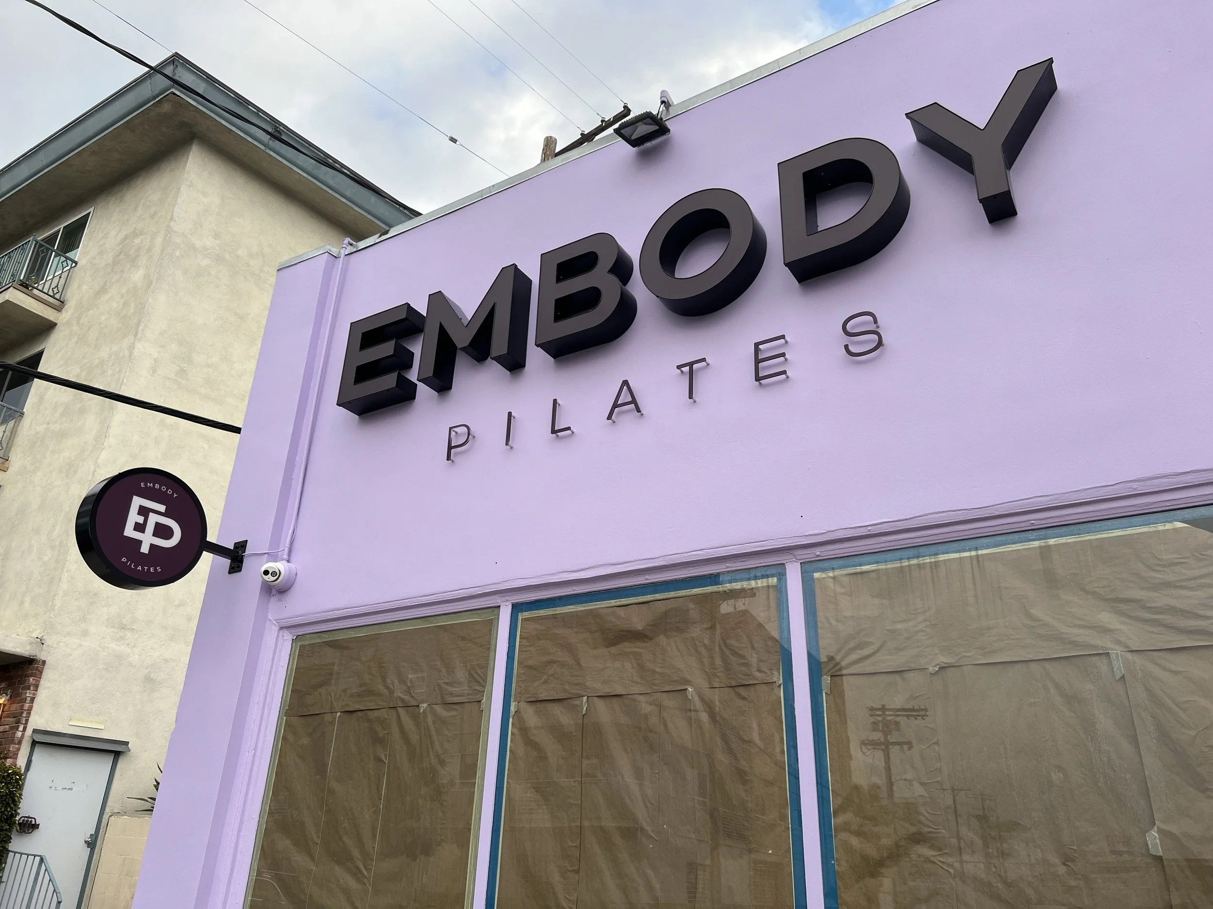 Sign for Empower Pilates in black 3D letters on a purple building exterior with a side round sign and large windows | Grizzly Signs | Los Angeles, CA | Custom Signs