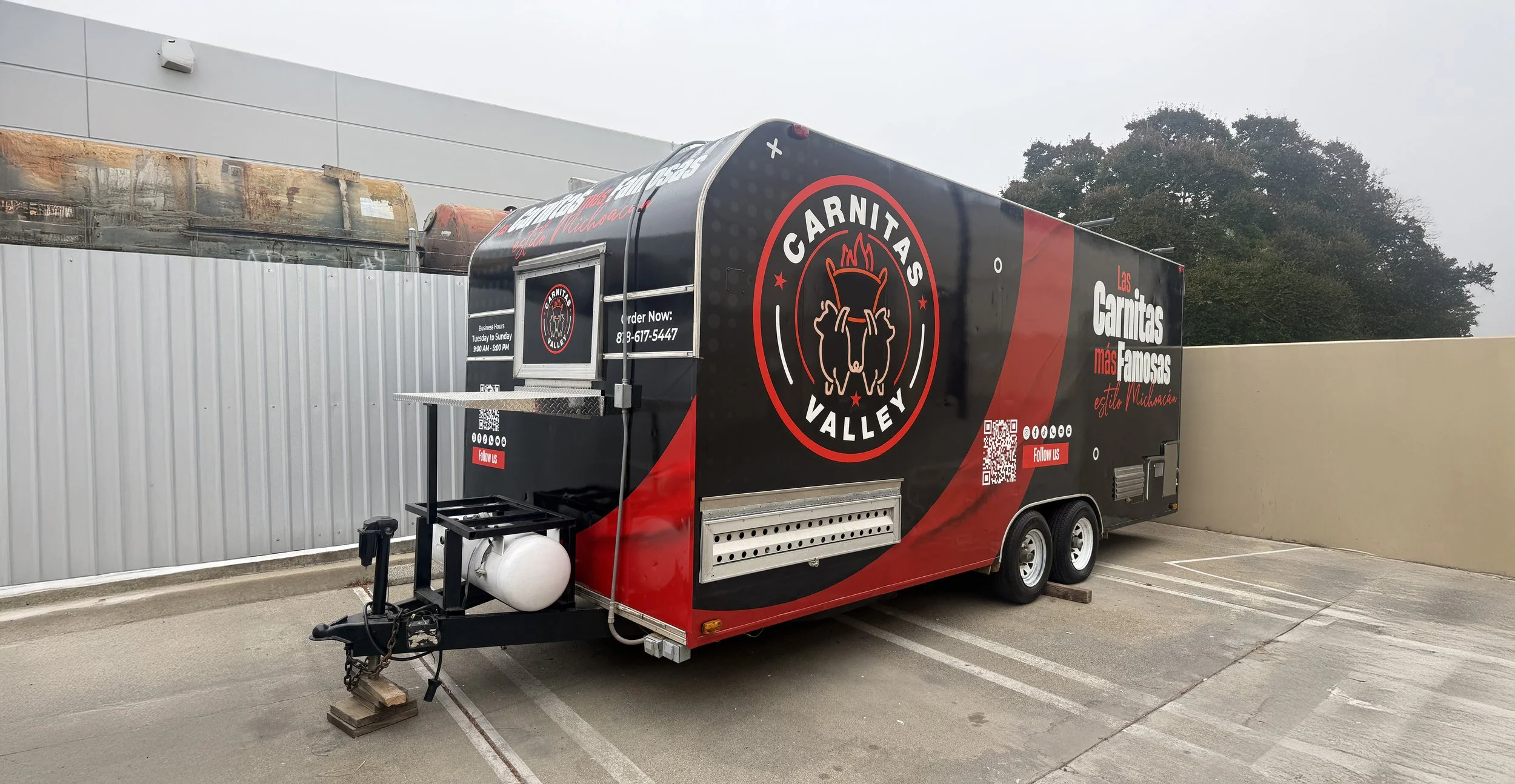 Black and red food truck with the logo 'Las Carnitas Valley' and text promoting Mexican cuisine, parked in a lot near a beige wall and a gray building, with trees in the background | Grizzly Signs | Los Angeles, CA | Custom Wraps