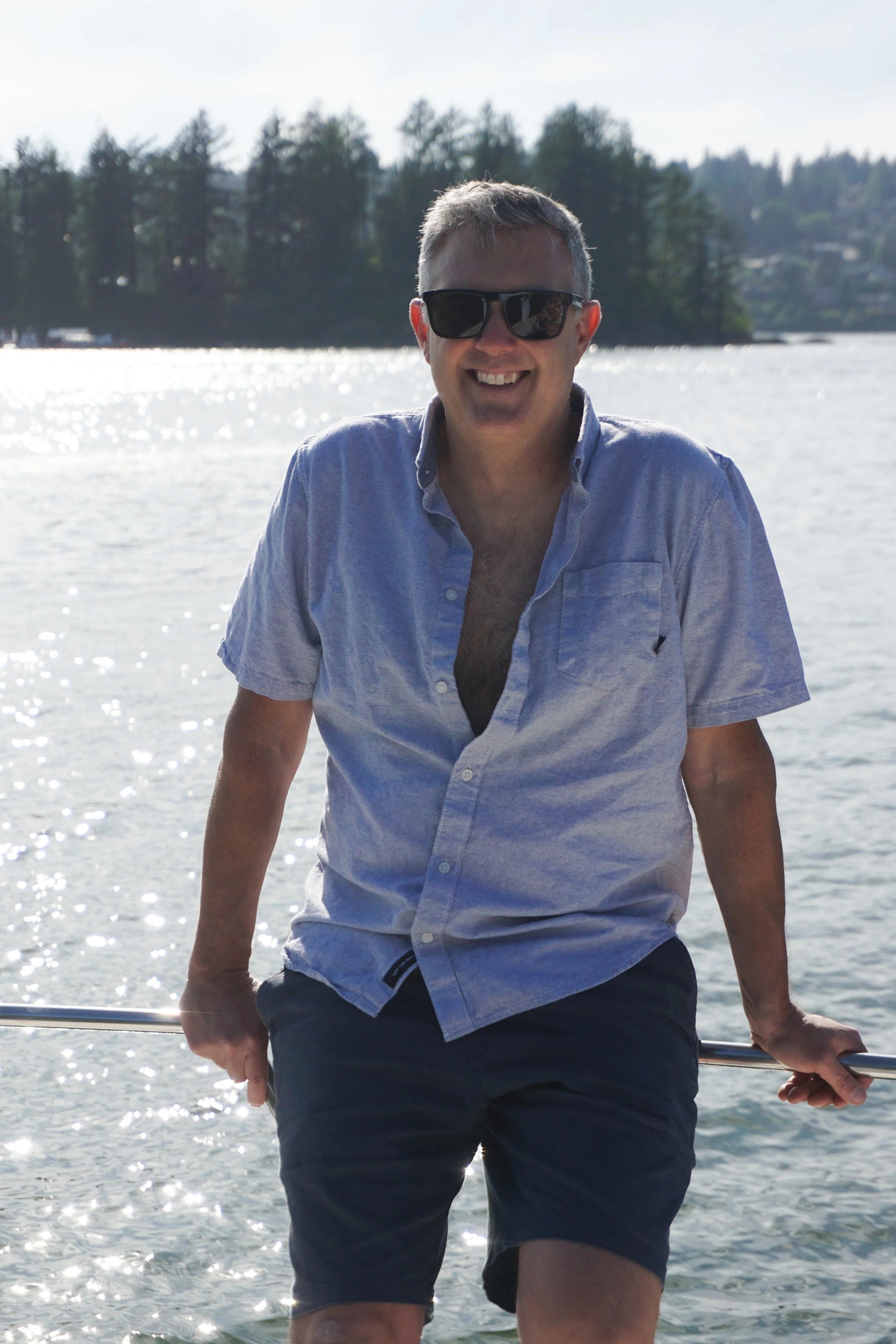 Smiling man wearing sunglasses and a light blue shirt, standing on a boat in front of a body of water with trees in the background.